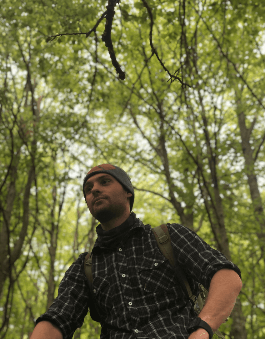 A man in a plaid shirt and beanie, carrying a backpack, stands in a lush green forest, looking off to the side. The dense canopy of trees creates a serene atmosphere.