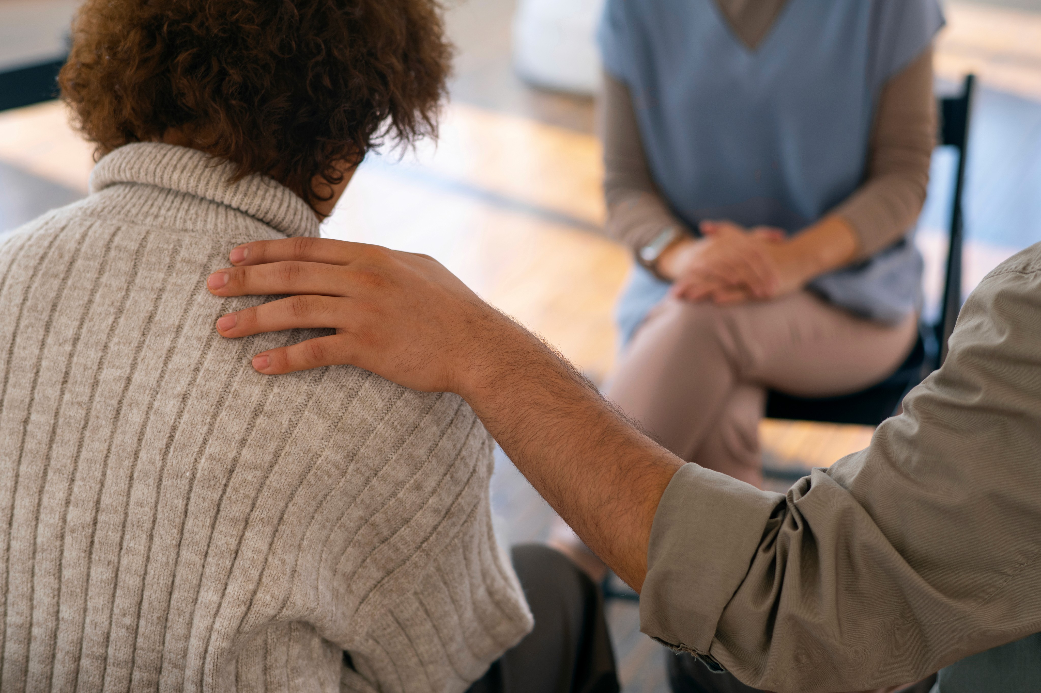 A person places a comforting hand on another person’s shoulder during a psychosocial recovery coaching, while a third person sits across from them.