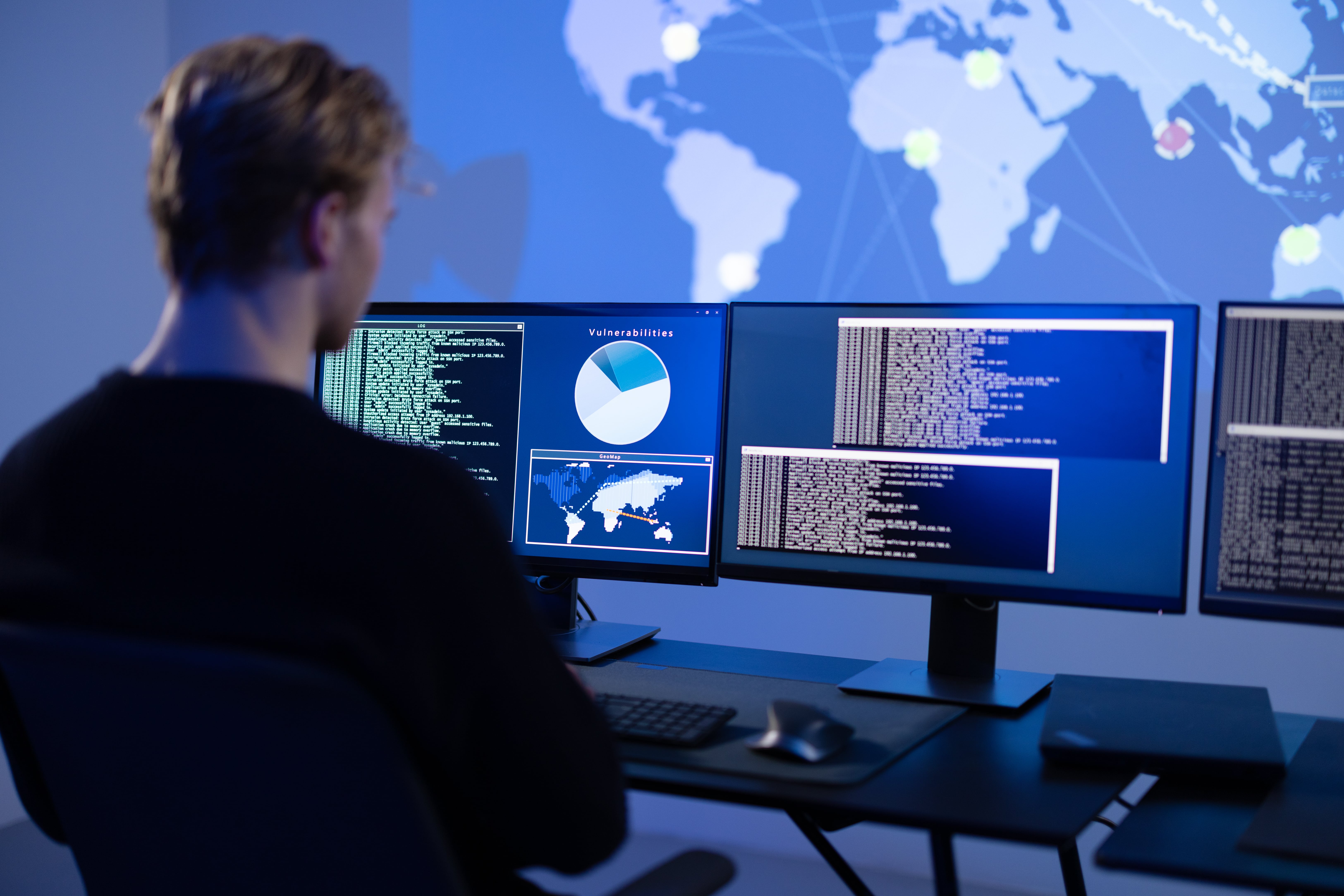 Cybersecurity analyst monitoring global threat data and code on multiple screens in control center