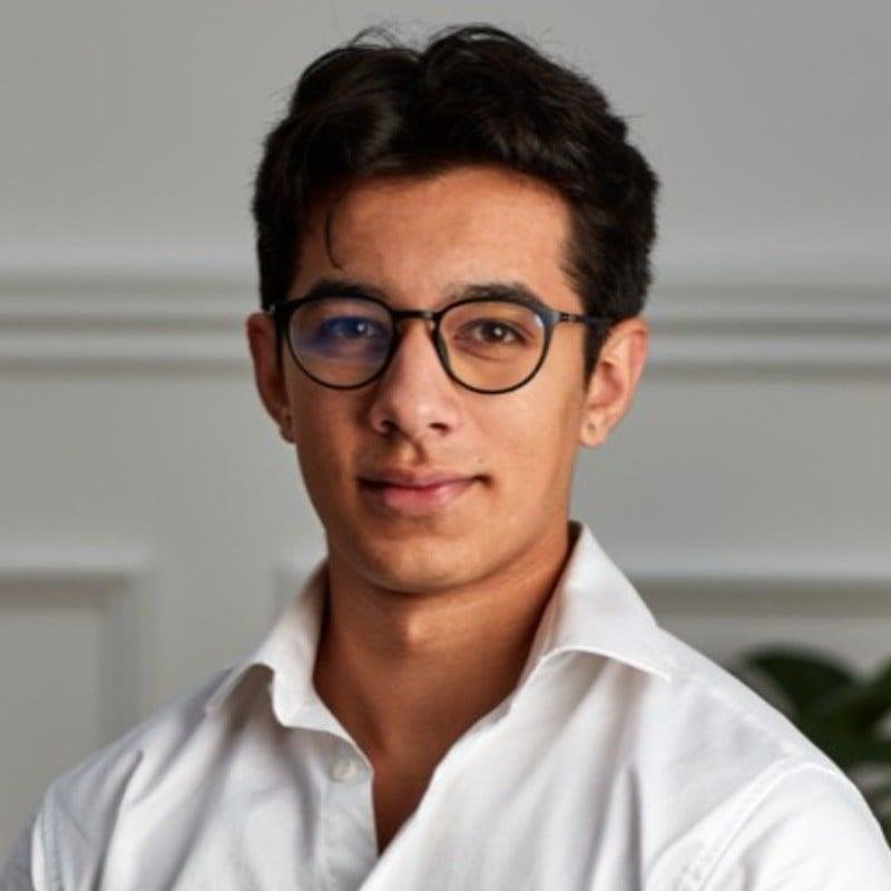 Man with brown hair wearing a black turtleneck and blazer, smiling slightly. The background is softly blurred, suggesting an outdoor, sunny setting.
