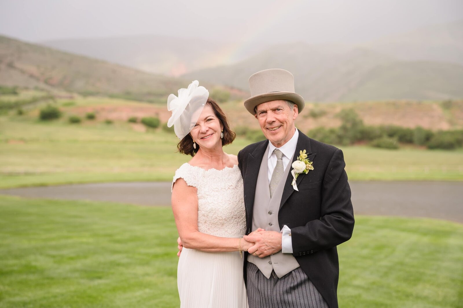 Couple in formal hats holding hands on a lawn beneath a faint rainbow