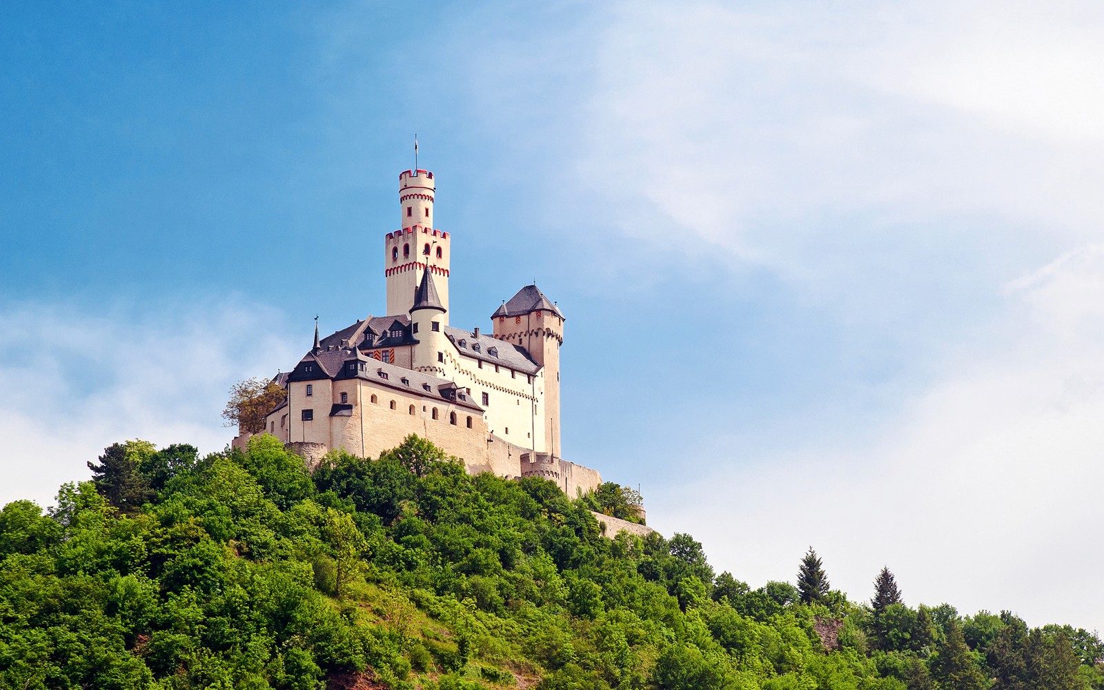 Marksburg Castle on a hilltop surrounded by lush greenery under a clear blue sky.