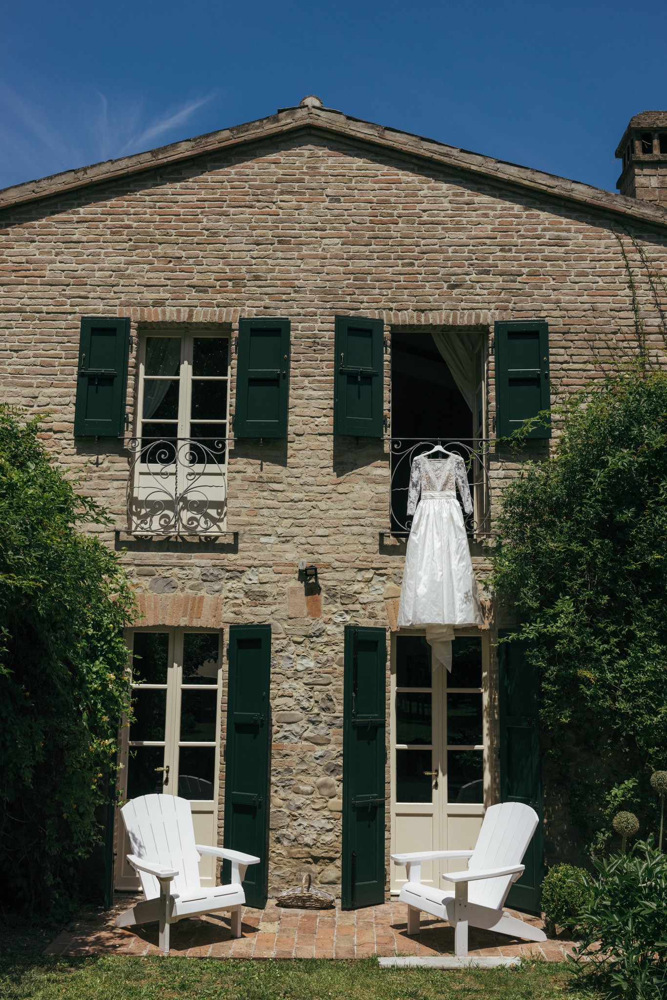 Abito da sposa sospeso alla finestra della villa, luce naturale su mattoni e persiane verdi, preparativi di matrimonio