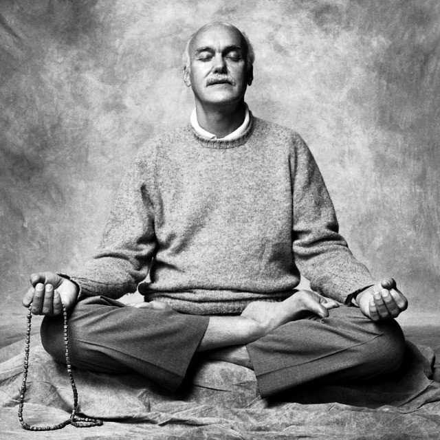Black-and-white portrait of a bald man meditating cross-legged while holding prayer beads.