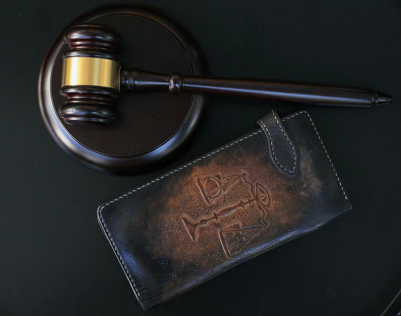 Um martelo de madeira escura com uma faixa dourada está colocado ao lado de uma pasta de couro desgastada com um símbolo jurídico em relevo, sobre um fundo preto, destacando temas de lei e justiça.