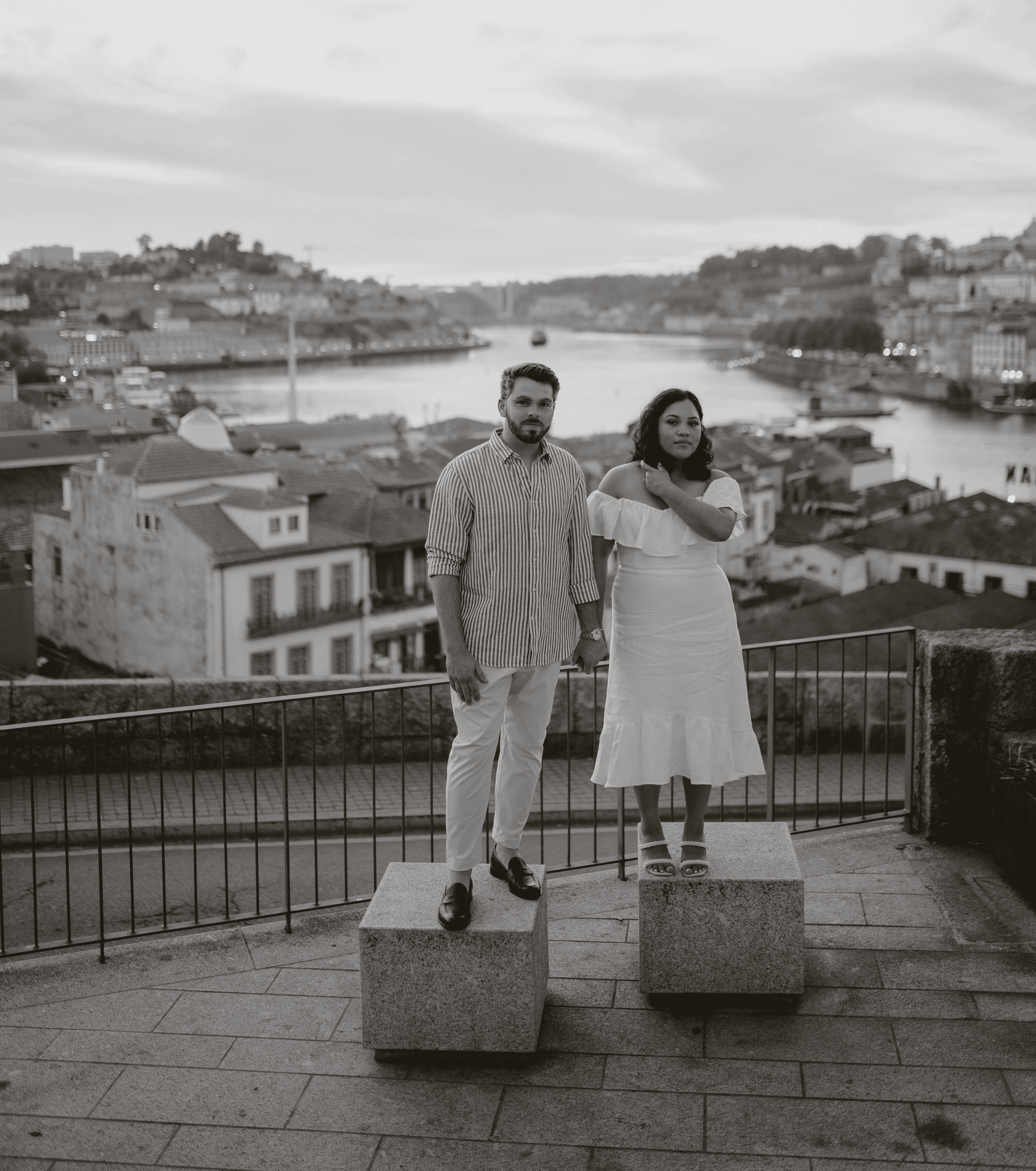 Black and white photo of a man and woman standing on stone blocks overlooking a cityscape and river. The man, dressed in a striped button-down shirt and light-colored pants, stands on the left. The woman, in an off-the-shoulder white dress, stands on the right with one hand on her shoulder. Behind them, a scenic view of a European city with buildings on hills and a river winding through the center is visible at sunset or twilight. A railing lines the overlook where they are standing.