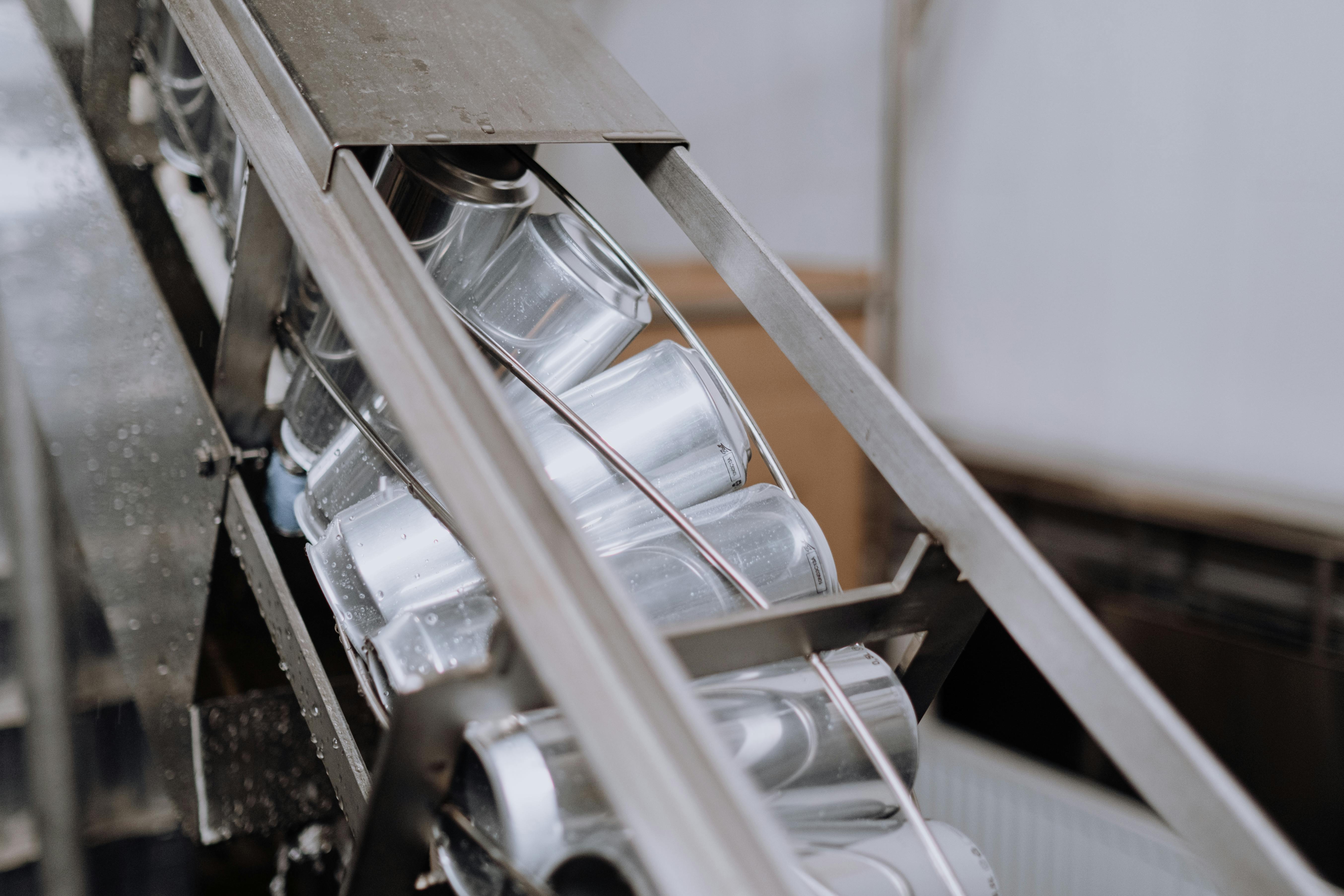 A close-up of a metal conveyor system in a manufacturing or packaging facility, with aluminium cans being being transported along the conveyor.
