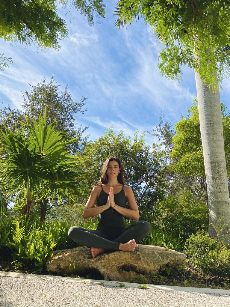 Woman seated in a meditative yoga pose outdoors on a rock with hands in prayer position wearing dark Treelance activewear