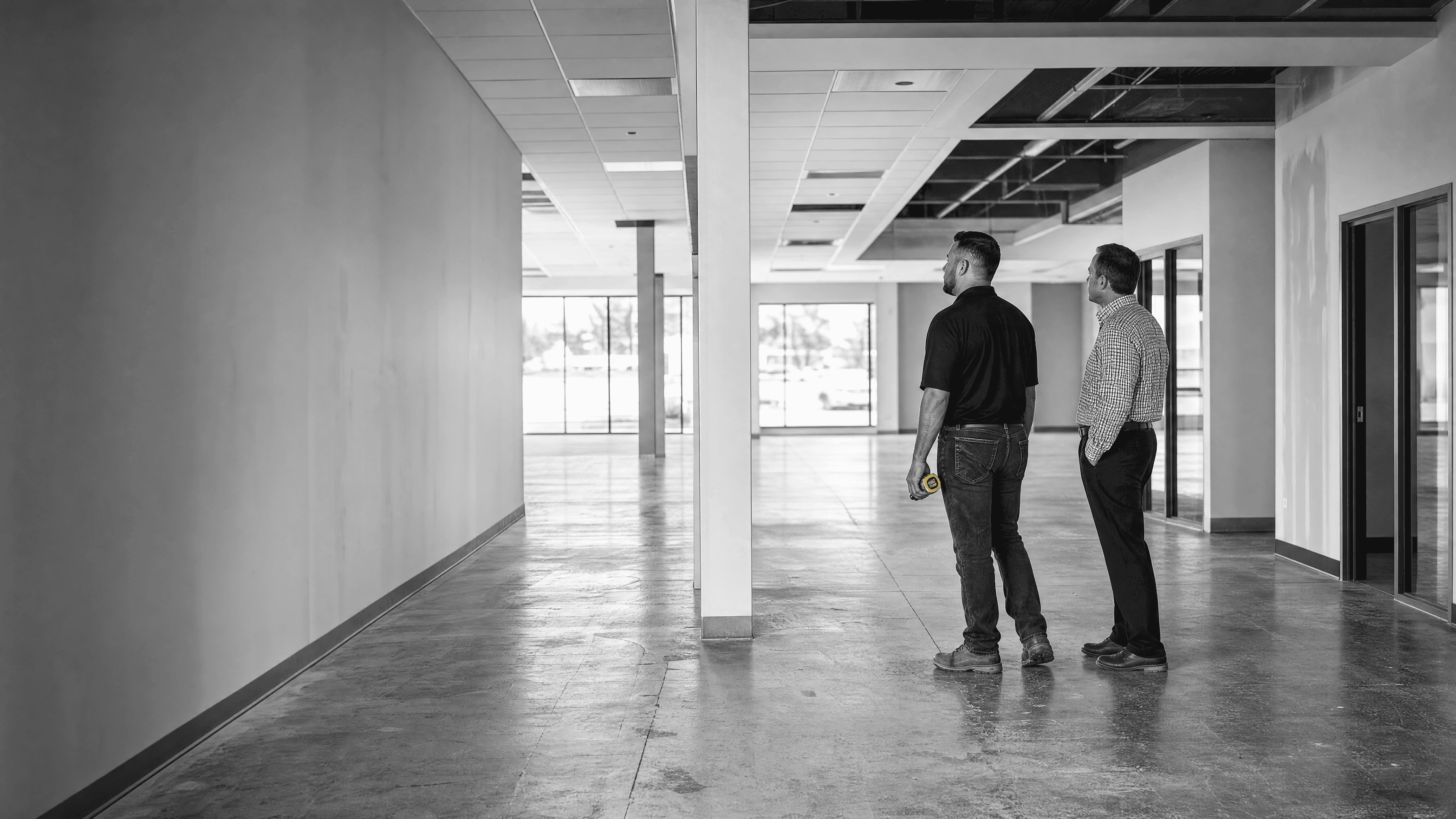 Two professionals reviewing an unfinished commercial interior with exposed ceiling areas and a tape measure.