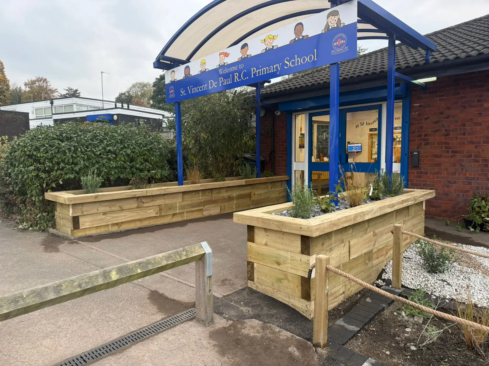 Wooden planter boxes with plants, situated near a blue and white awning entrance. Paved pathway visible.