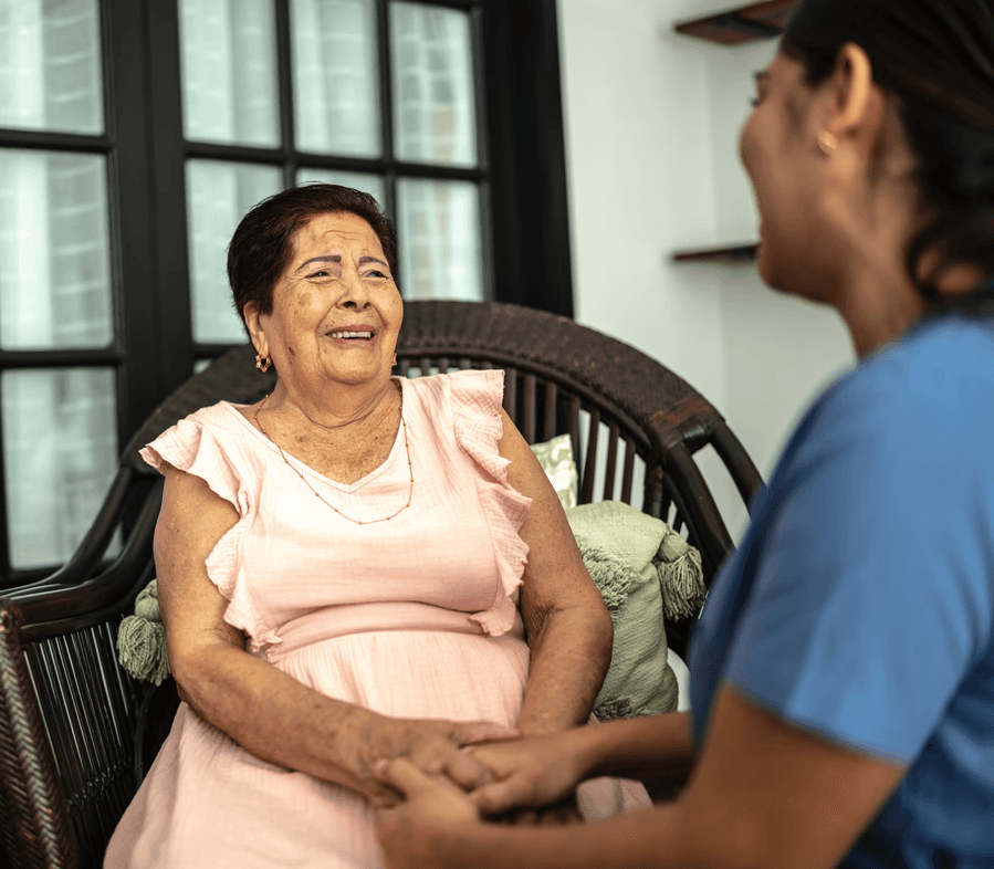Elderly woman in pink dress smiling while sitting in a wicker chair, holding hands with caregiver in blue uniform by a window