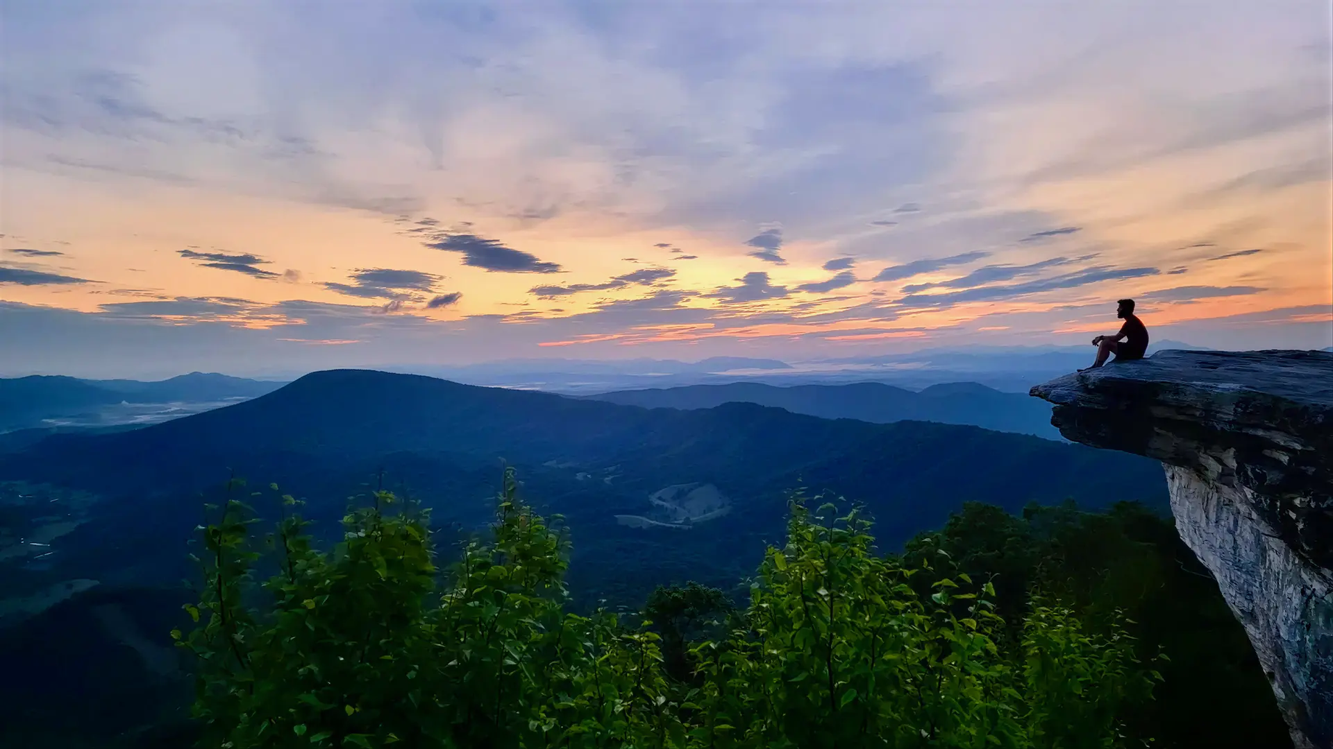 A person sitting on a rocky outcrop at dusk, overlooking layered mountain ridges beneath a wide sky.