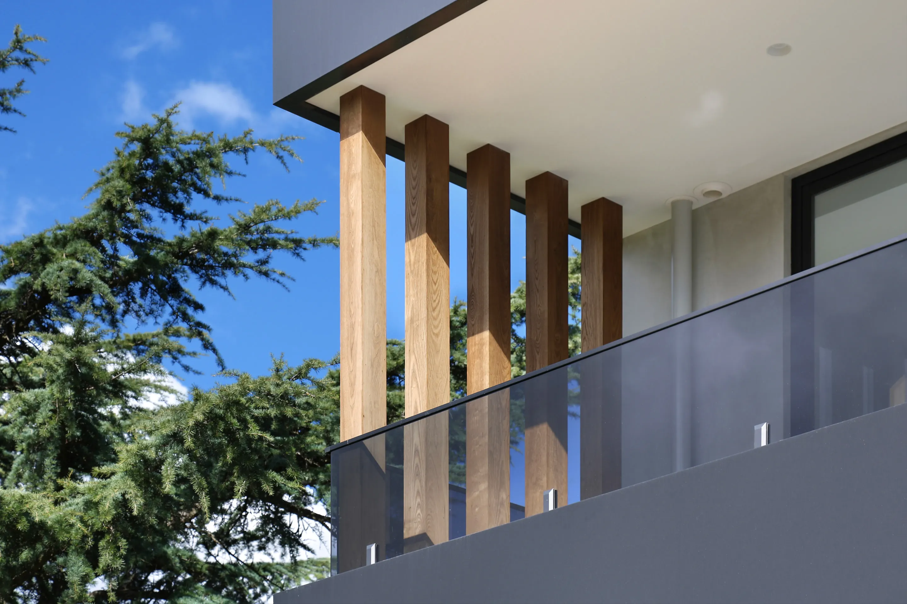 Detail of the upper balcony with five square timber posts, a glass balustrade and a charcoal-clad soffit against blue sky and a tall conifer
