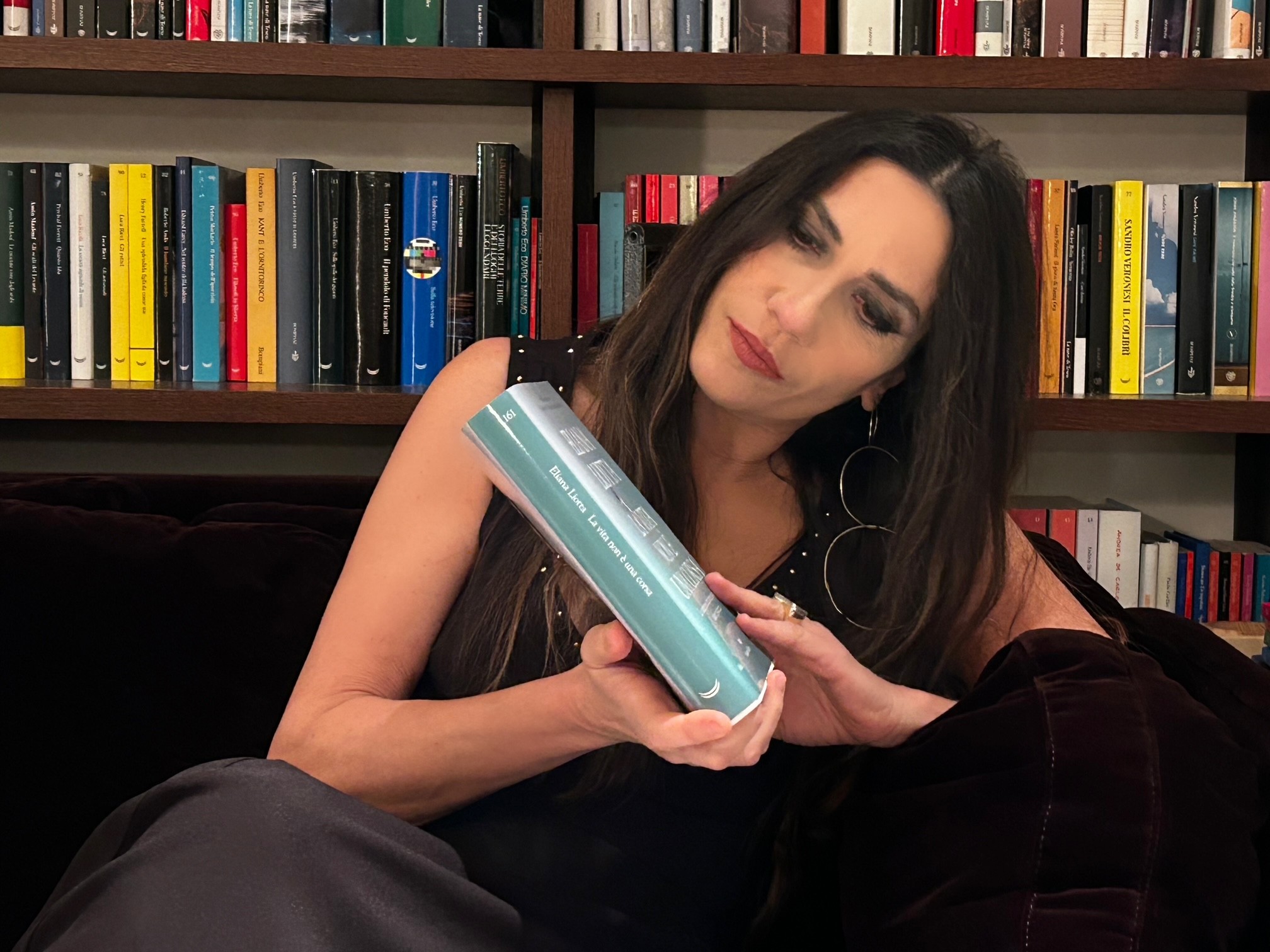 A young person with curly hair smiles at the camera while sitting in front of a laptop against a dark background.