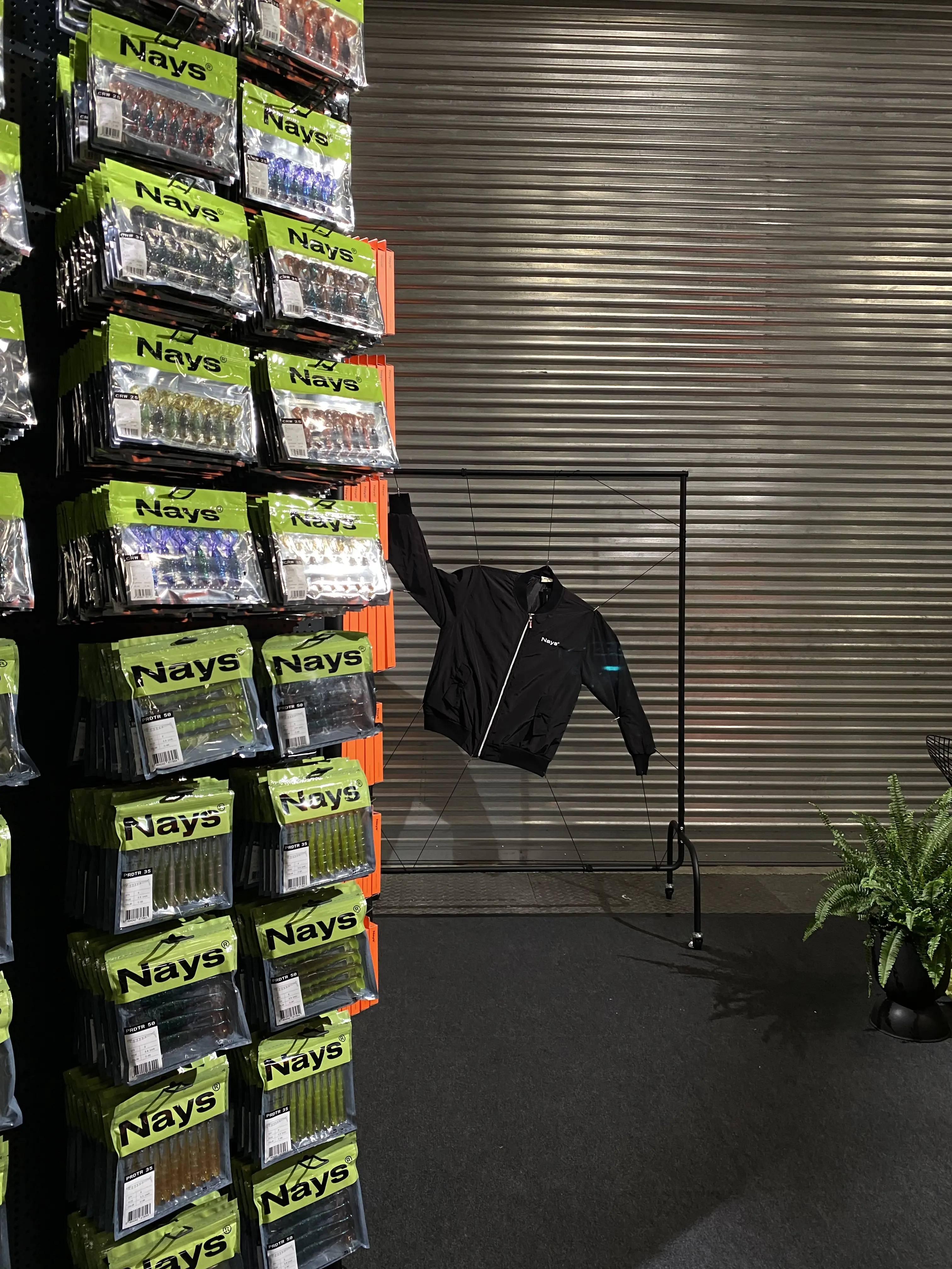 Display tower stacked with neon-green Nays lure packaging next to a black jacket on a clothes rail.