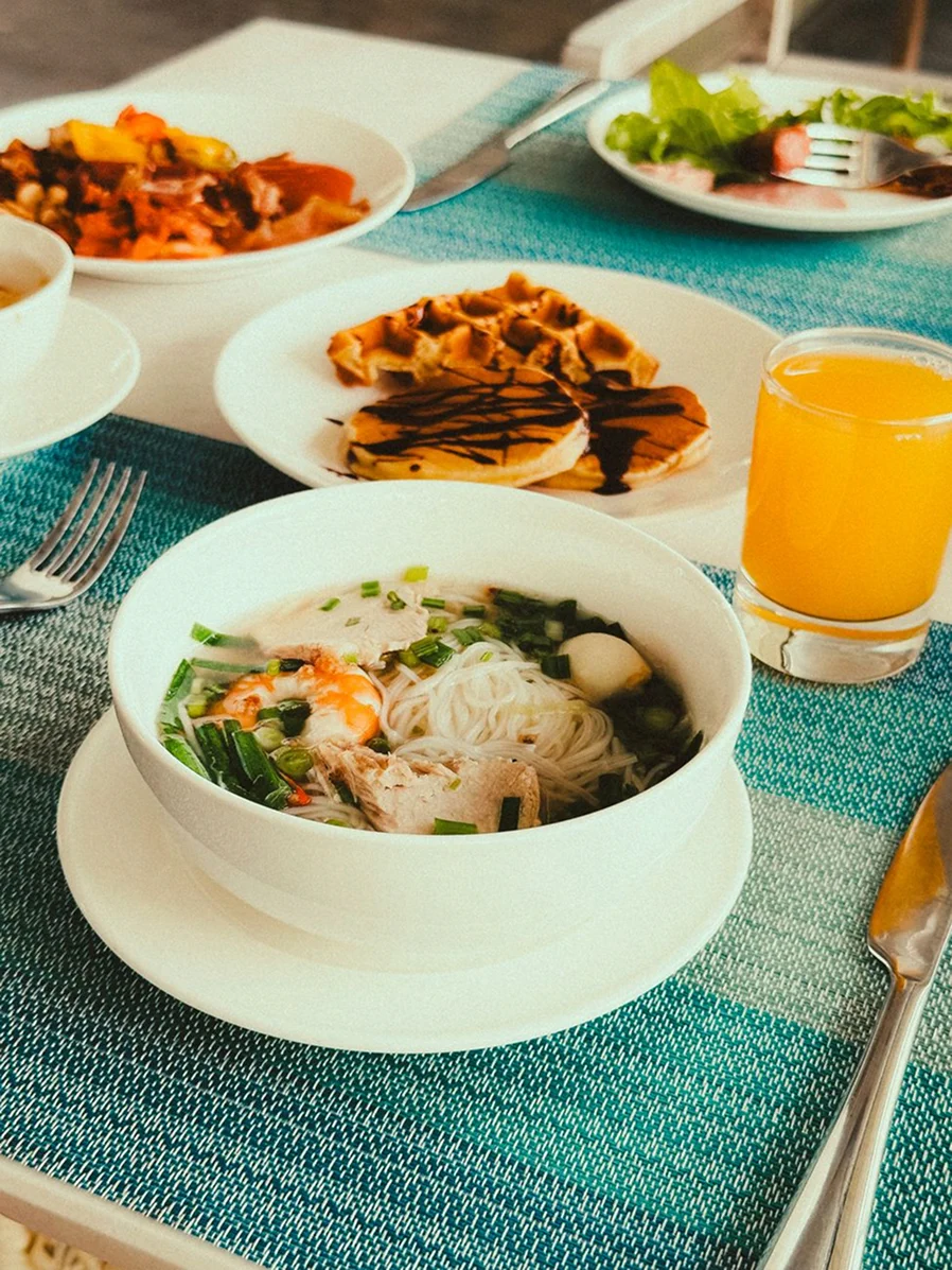 Casual resort breakfast table with noodles, waffles, side dishes, and fresh orange juice.
