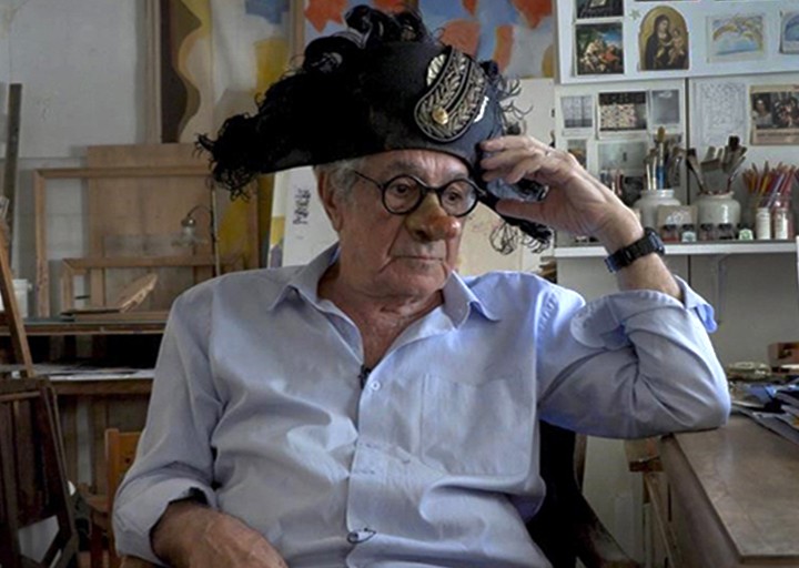 Ο Αλέξης Κυριτσόπουλος, με καπέλο του Ναπολέοντα και μάσκα, καθισμένος στο εργαστήριό του ανάμεσα σε έργα και σχέδια