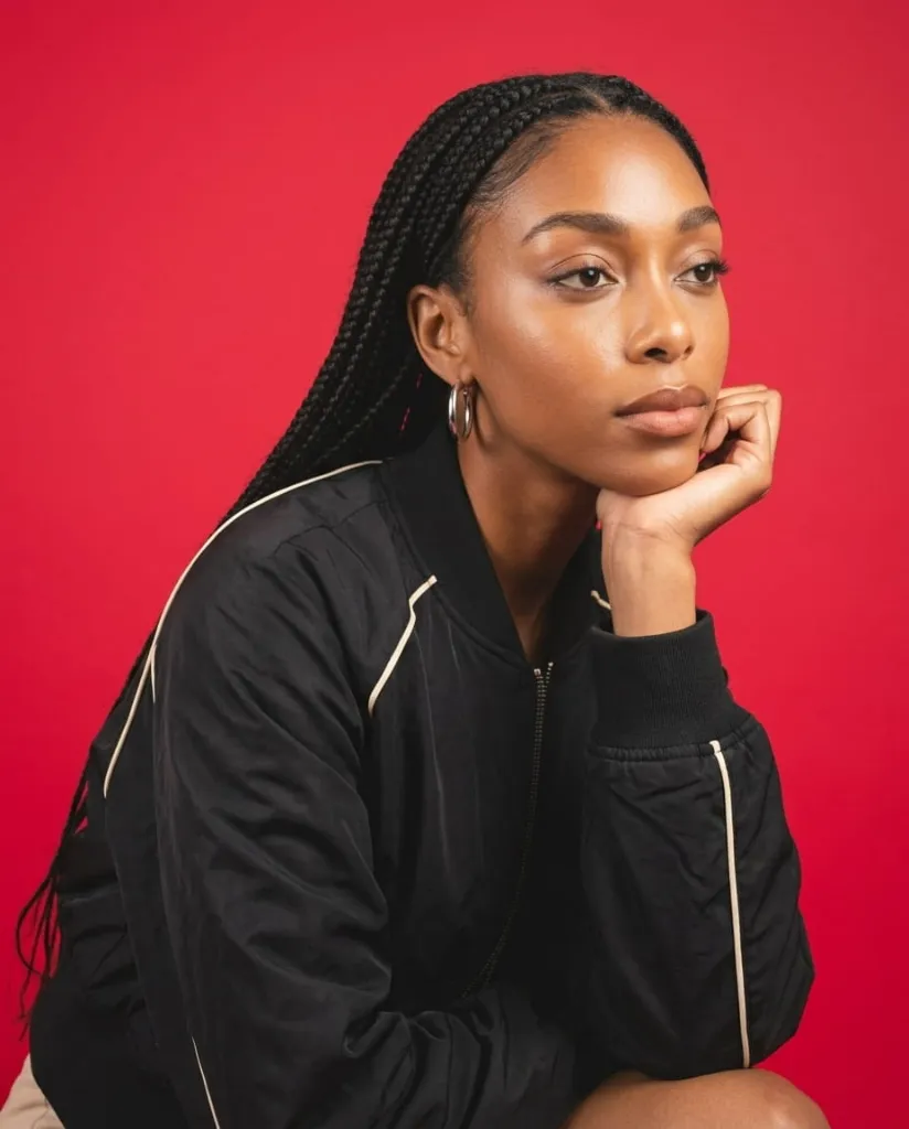 A young woman with braids and a black jacket poses against a red background.