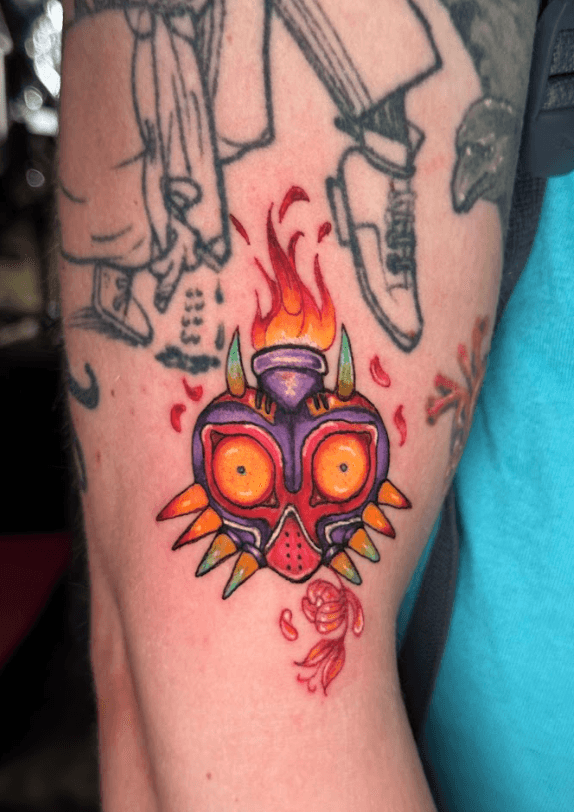 A vibrant tattoo of a chainsaw-like creature on a person's forearm. The tattoo is detailed with shades of orange, black, teal, and red.