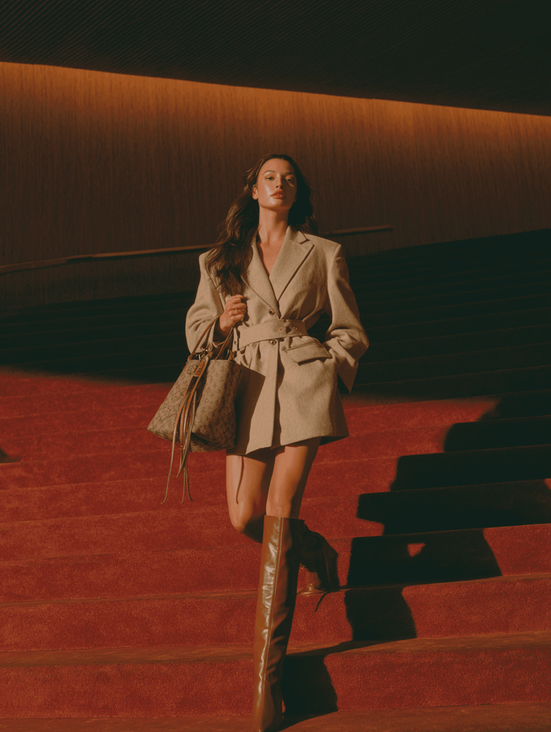 A stylish woman in a beige coat and brown boots descends a red-carpeted staircase, exuding confidence and elegance.