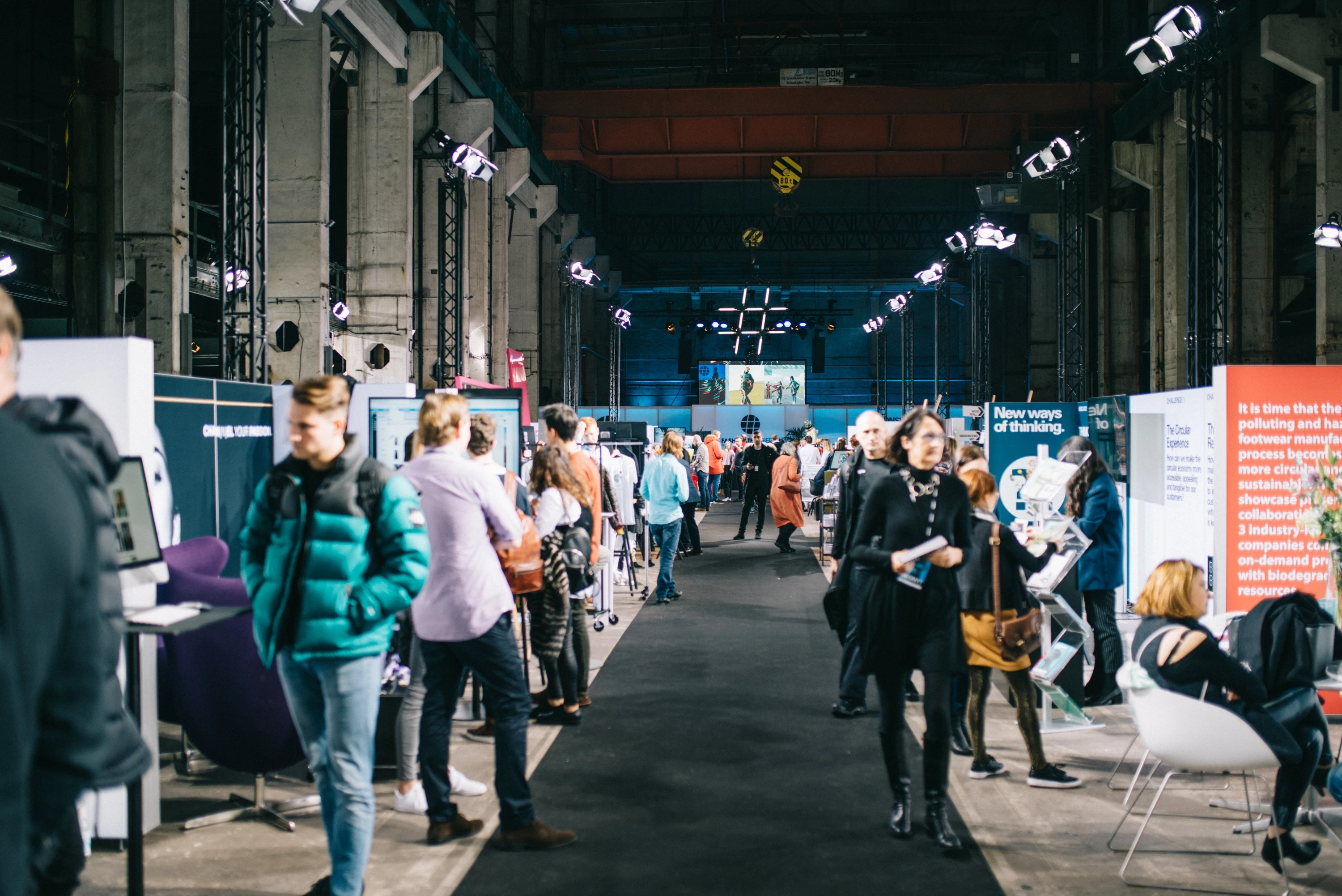 Visitors strolling between exhibitor stands and presentation walls in the industrial hall at FASHIONTECH Berlin.