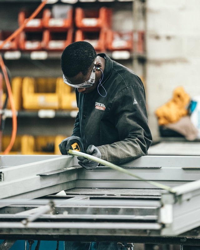 Factory worker wearing safety glasses using a tape measure on a piece of metal.