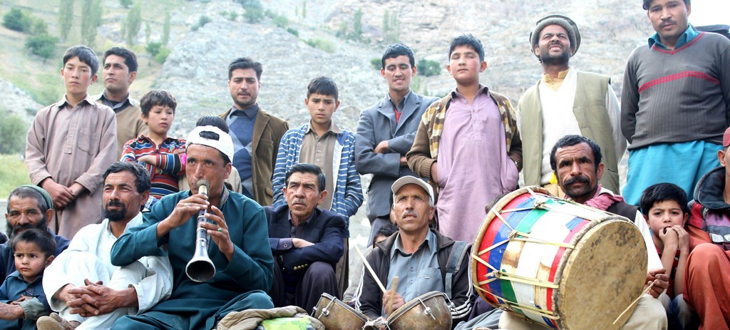 A group of men and boys in traditional attire gather outdoors, with musicians playing instruments such as a drum and a flute, capturing a vibrant cultural celebration in a mountainous region.