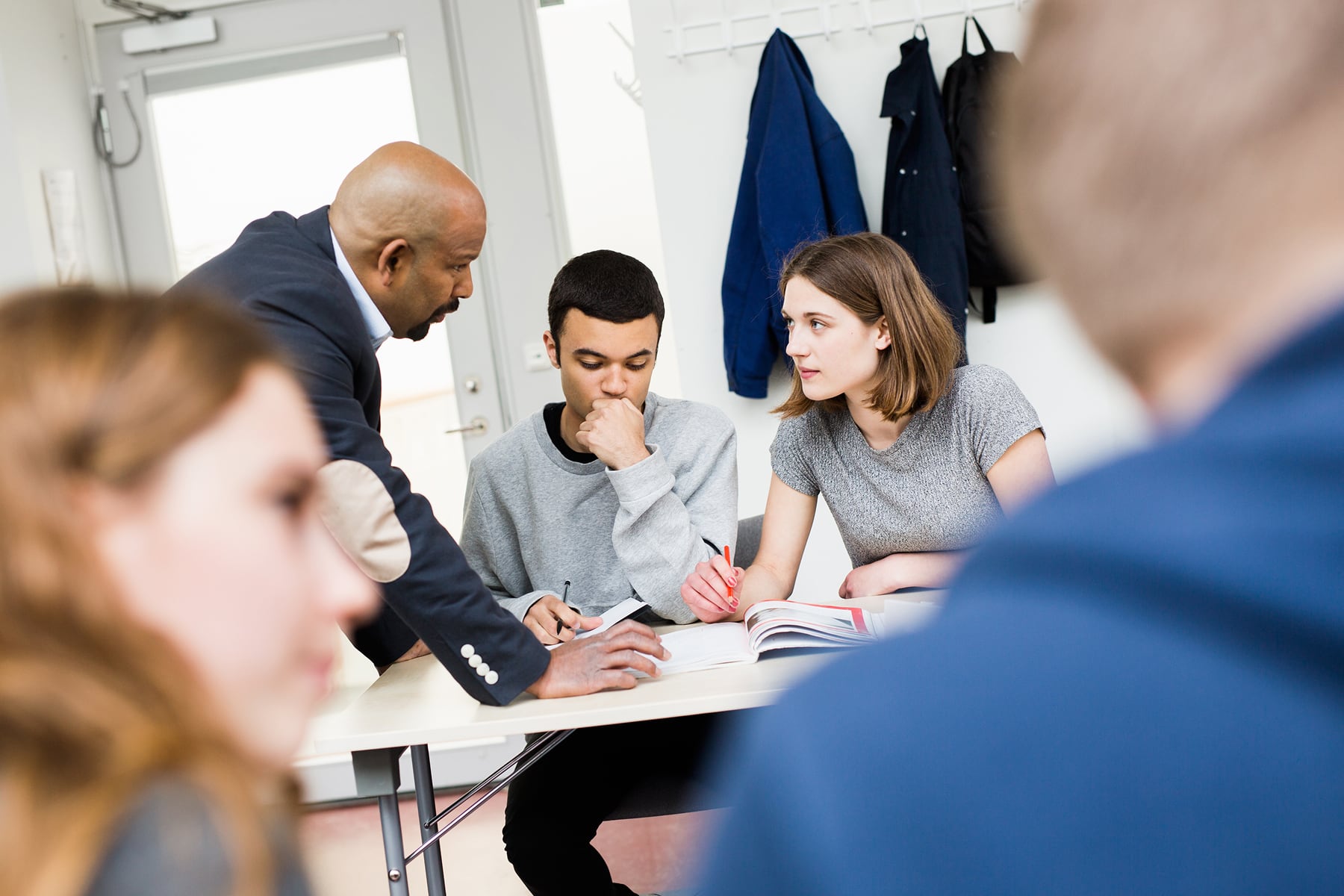 A teacher leans over a table to support two students who are studying together, with other classmates blurred in the foreground.