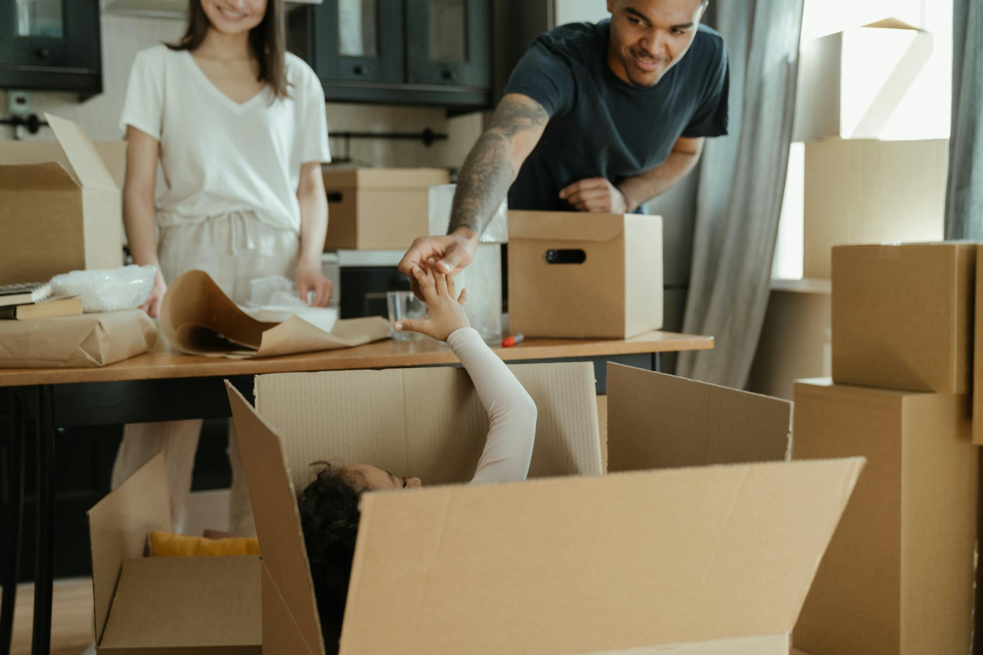 A lighthearted scene of a family during a move. A man is leaning over a large, open cardboard box and playfully holding the hand of a child lying inside it. A smiling woman is standing in the background next to a table covered with packing paper and other moving supplies, with many stacked boxes visible throughout the room.