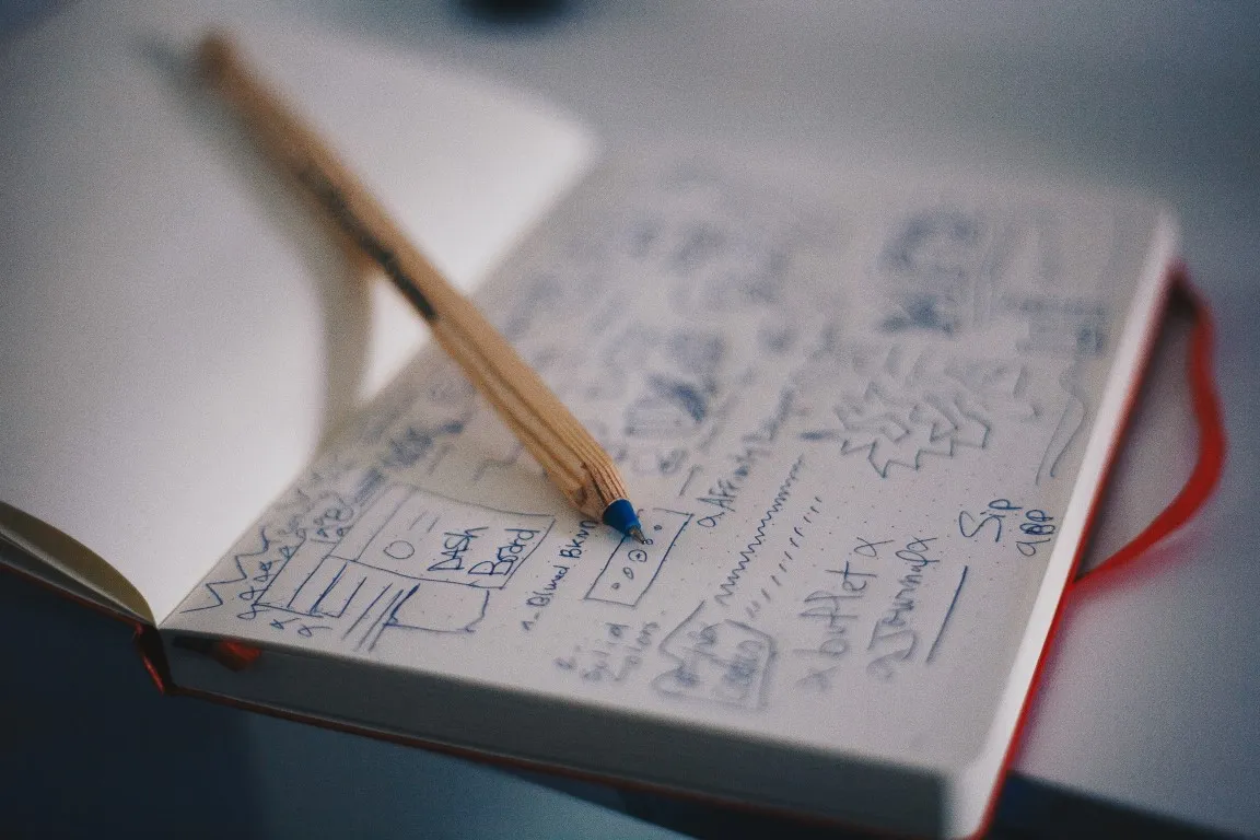 A wooden pencil with a blue tip rests on an open notebook filled with handwritten notes, sketches, and diagrams, possibly representing brainstorming, planning, or creative ideation for design or development.