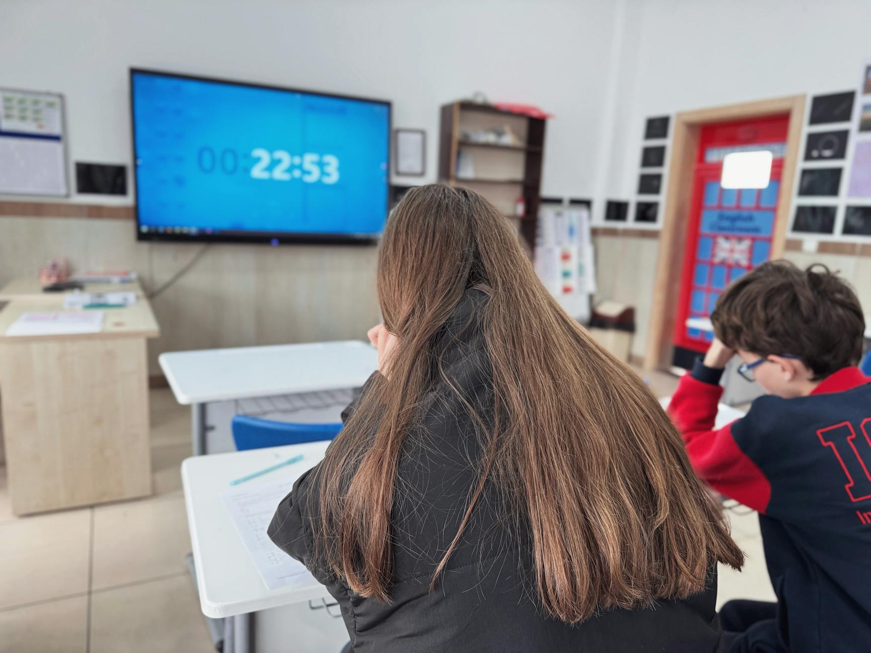 A teacher pointing to a colorful stopwatch on a digital whiteboard while students sit ready at their desks.