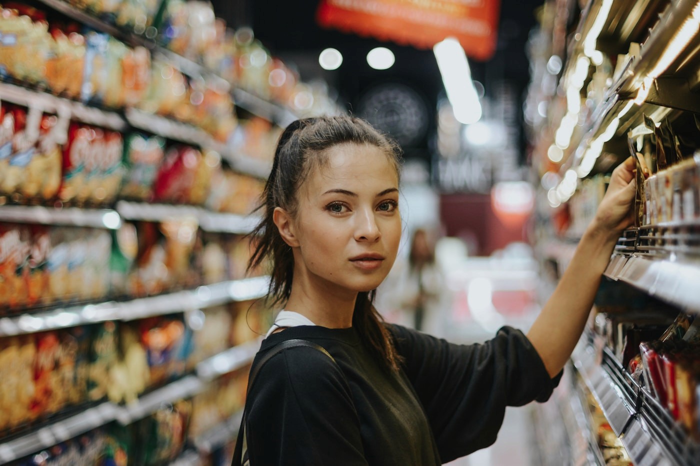 A woman reading a product package in a grocery store aisle, checking the ingredient and Contains label for allergens