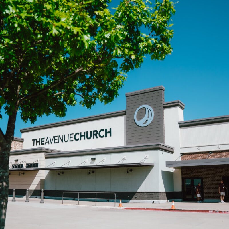 A modern church building with a large sign, surrounded by a clear blue sky and green trees nearby.