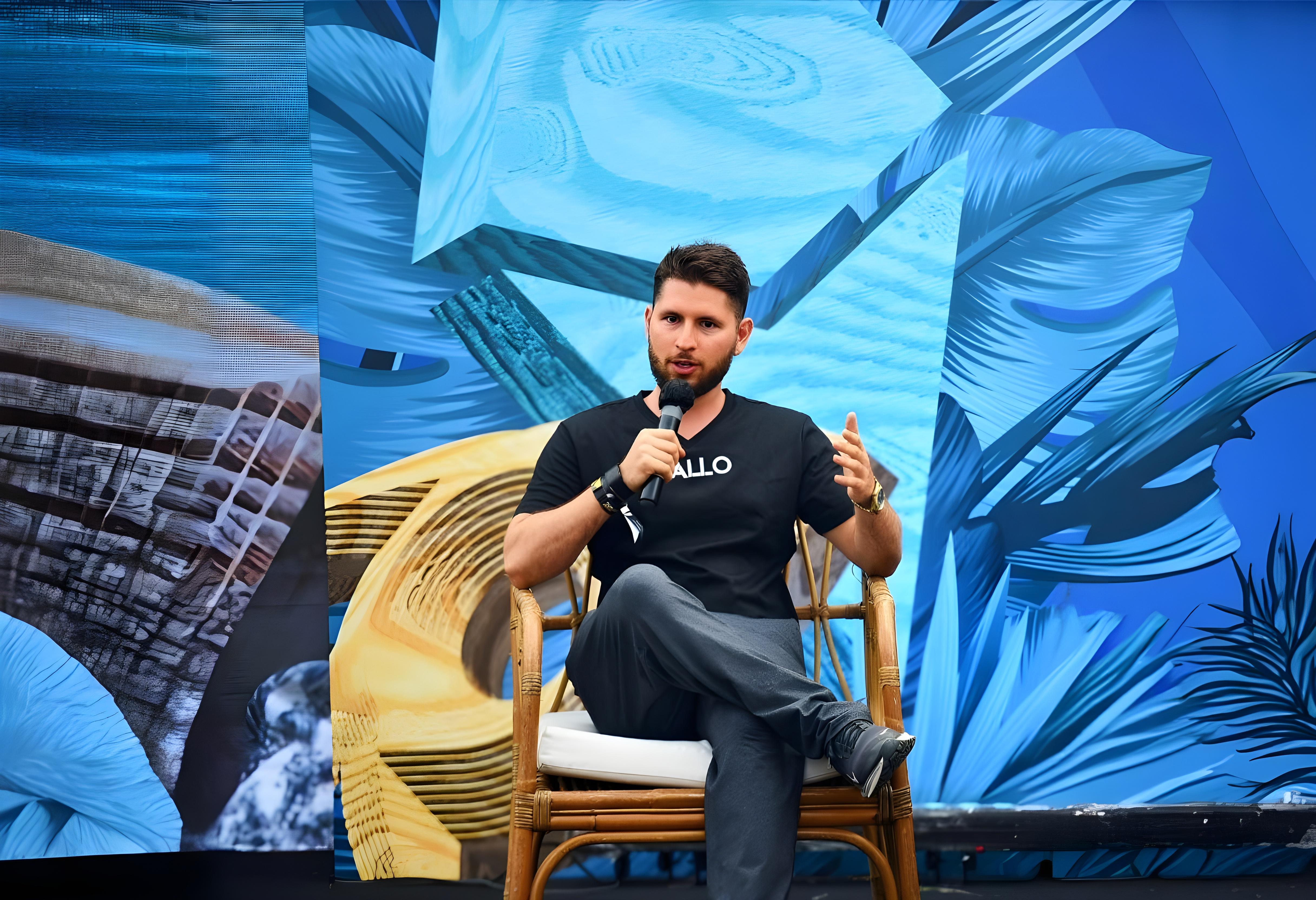 Kingsley, CEO of Allo.xyz, speaks at an event, passionately discussing blockchain innovation and real-world asset tokenization. Seated in a rattan chair, he wears a black Allo-branded T-shirt, gesturing expressively with one hand while holding a microphone in the other. Behind him, a striking blue abstract mural with geometric and organic patterns creates a futuristic atmosphere, reflecting Allo's vision for the future of decentralized finance.