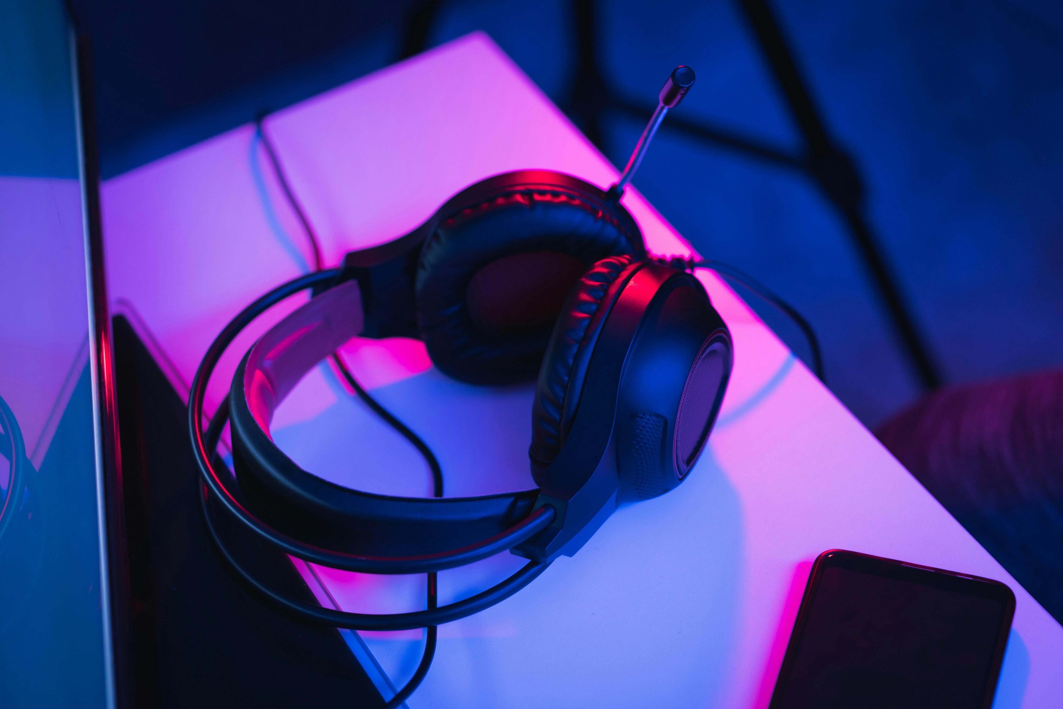 Black gaming headset with a microphone resting on a white desk, illuminated by blue and pink neon lighting.