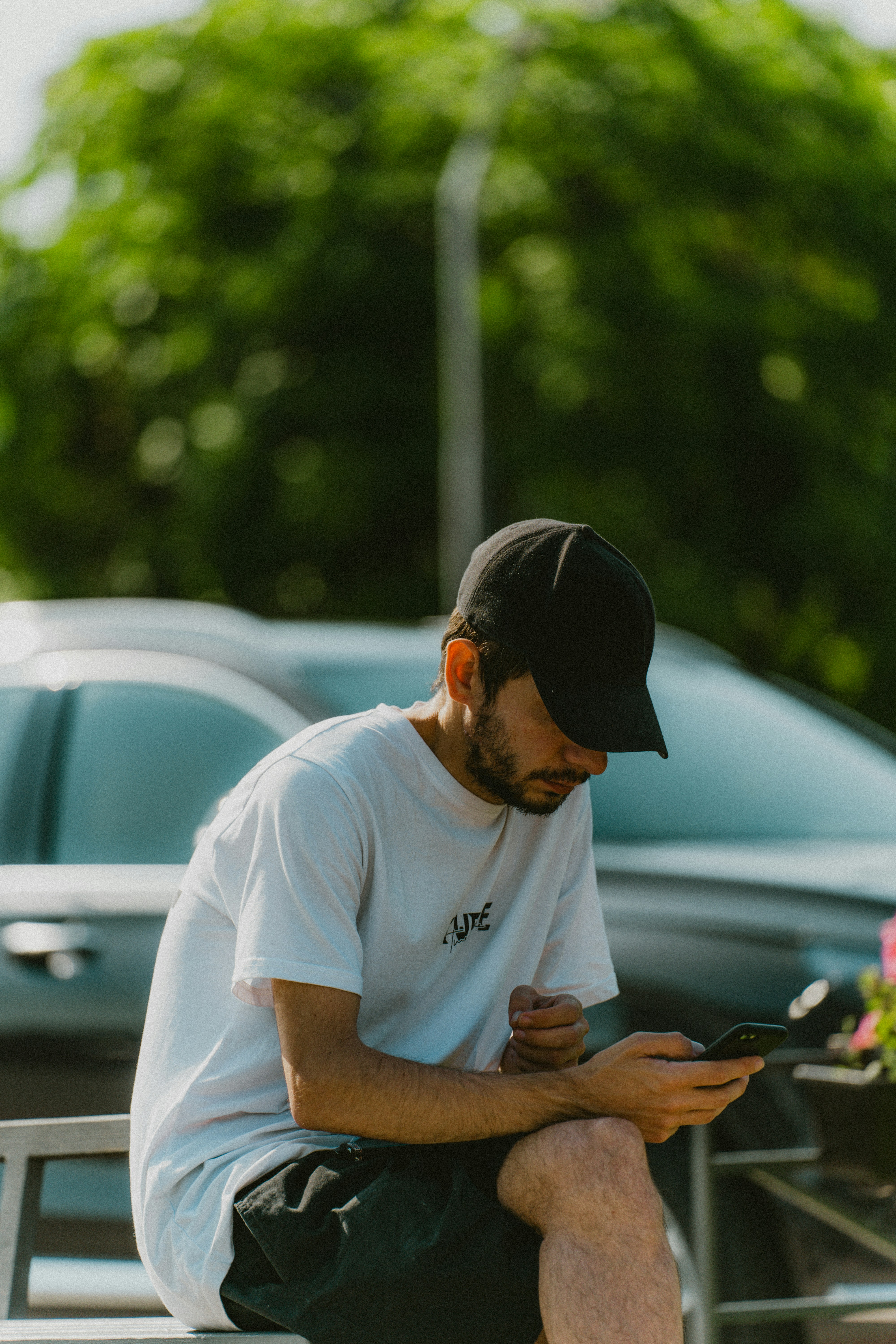A person in a white t-shirt sits on a car, looking at their phone, with greenery in the background.