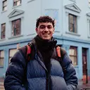 Person wearing a blue puffer jacket and red backpack standing in front of a light blue building on a city street.