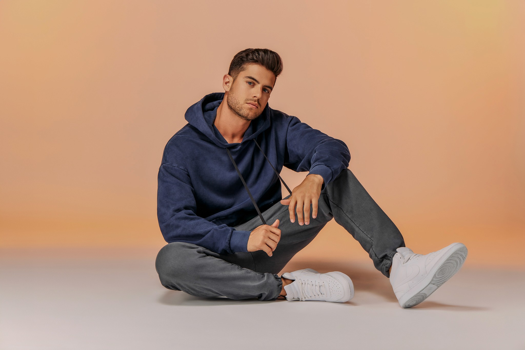 A person with short hair is sitting on the floor against a peach background, wearing a dark blue hoodie, gray pants, and white sneakers, holding the strings of their hoodie with one hand.
