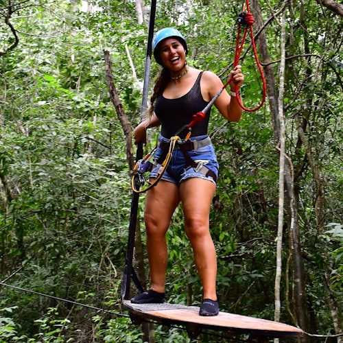 一位戴着头盔和安全带的女士站在木制平台上，在郁郁葱葱的森林中微笑，准备进行滑索活动。