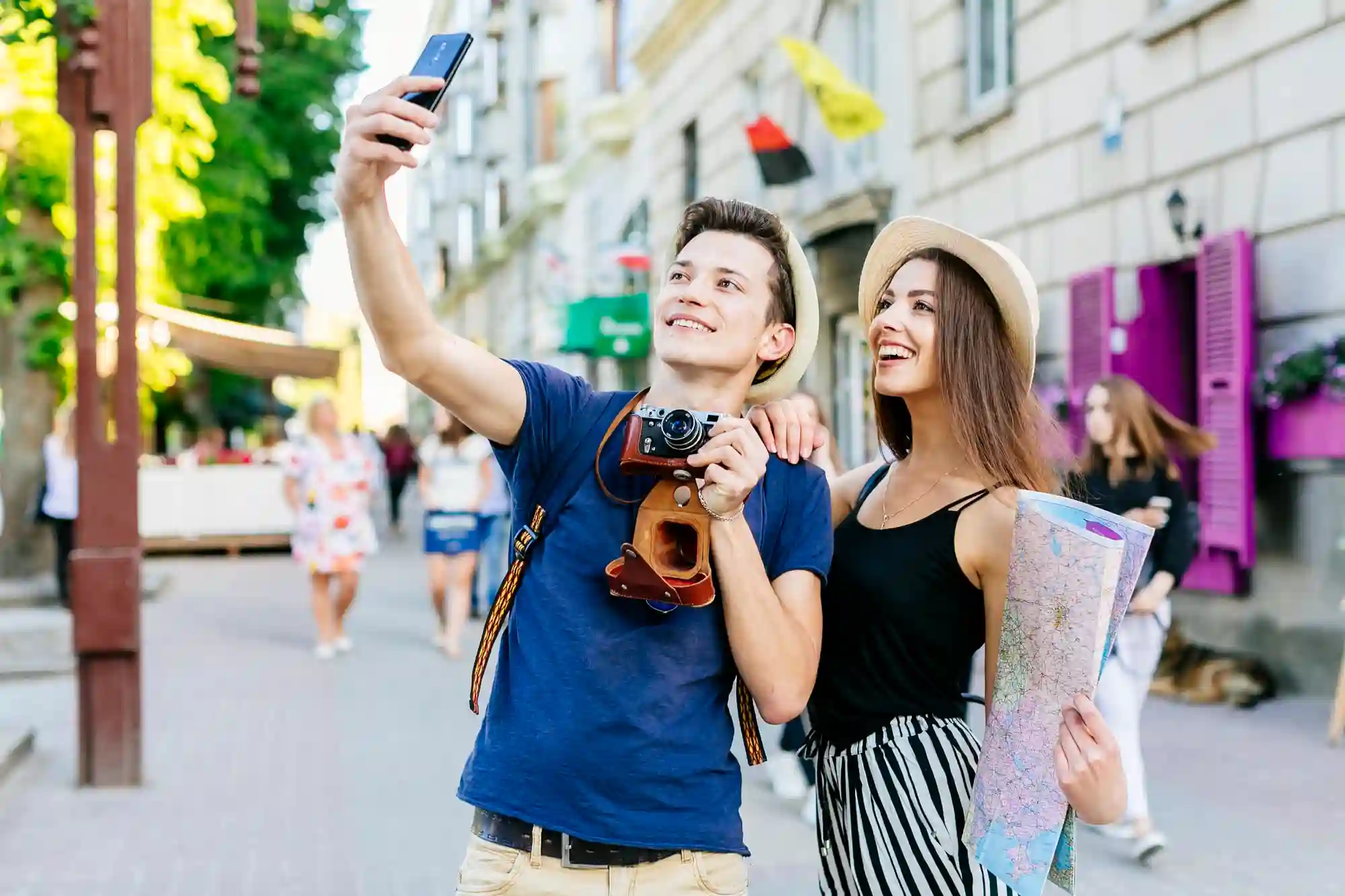 A happy young couple smiling and taking a selfie while exploring a sunny, European-style city street with a map.