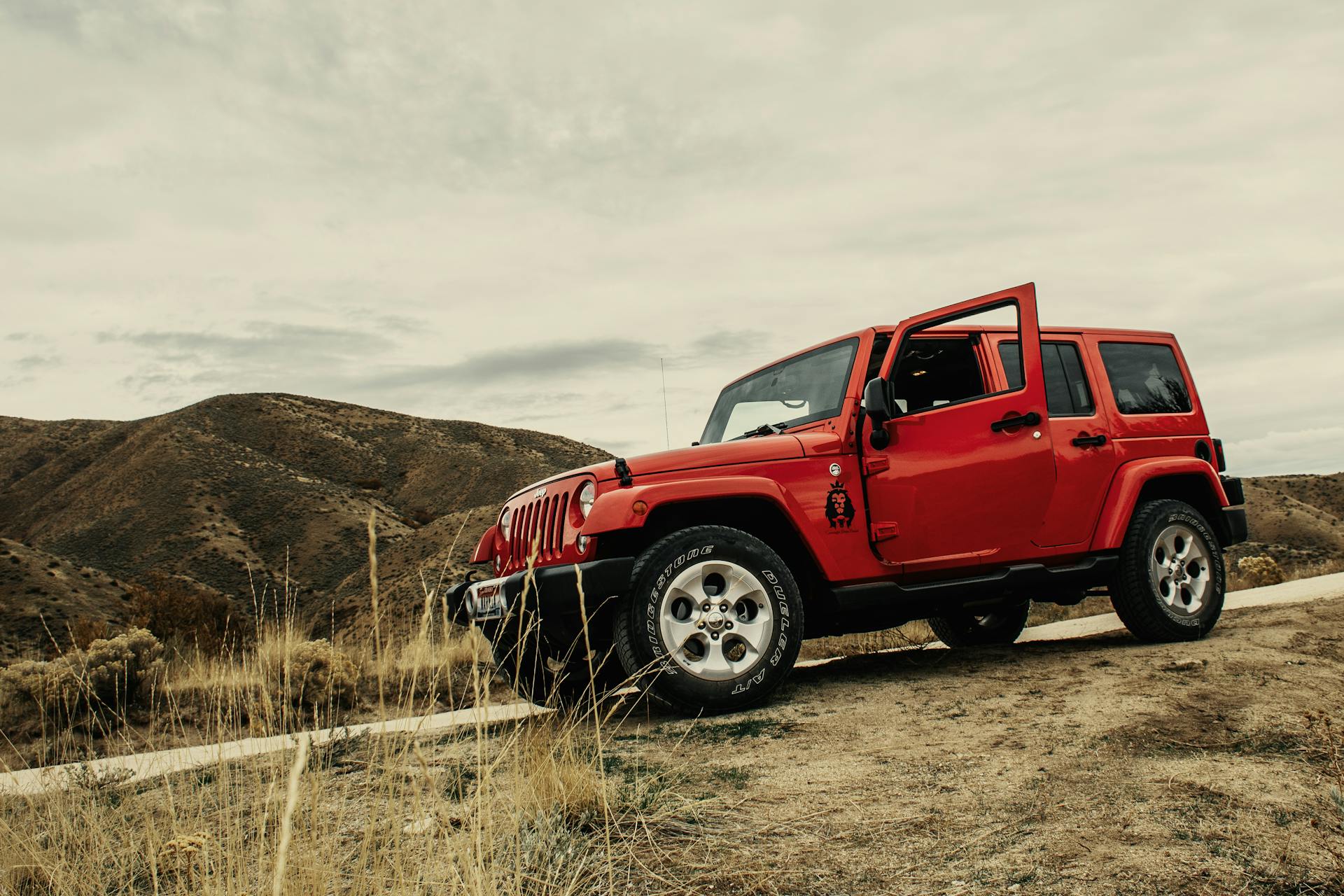A bright orange Jeep Wrangler, with the driver's side door open, parked on a dirt road in a dry, hilly landscape. The sky is overcast.