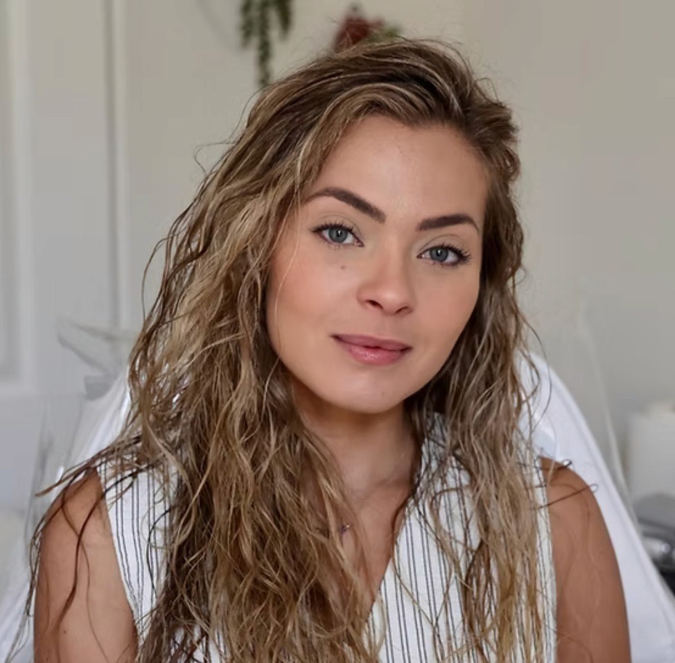 Young woman with wavy blonde hair and a gentle smile sits indoors against a blurred background. She wears a white striped top and exudes a calm, warm demeanor.