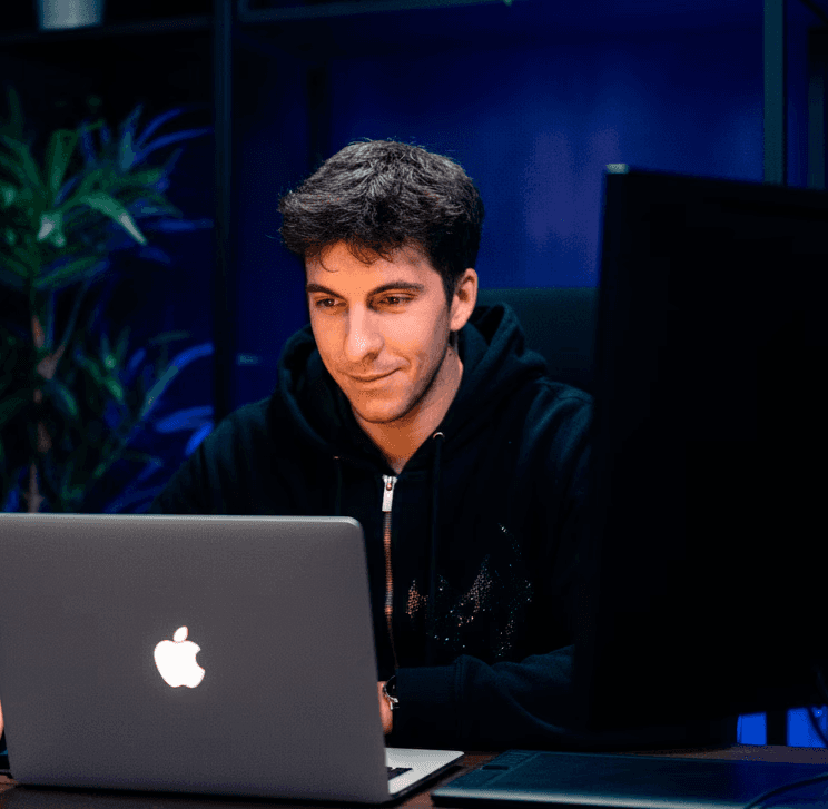 Jeune homme concentré, type créateur ou développeur, travaillant sur un ordinateur portable MacBook Pro argenté, posé sur un bureau en bois sombre. Il porte un sweat à capuche noir avec un zip ouvert au col. Son regard est dirigé vers l'écran de l'ordinateur portable, illuminant légèrement son visage. À sa droite, on distingue une tablette graphique avec son stylet, suggérant un travail de design ou de retouche. Un second écran d'ordinateur, éteint ou en veille, est visible à droite. L'arrière-plan est moderne, avec une étagère en métal noir contenant des plantes vertes luxuriantes (comme des pothos ou des philodendrons) et des éléments décoratifs sombres. L'éclairage ambiant est faible, créant une atmosphère studieuse et créative, avec des touches de lumière bleue profonde provenant de l'arrière-plan, éclairant le mur et l'étagère.