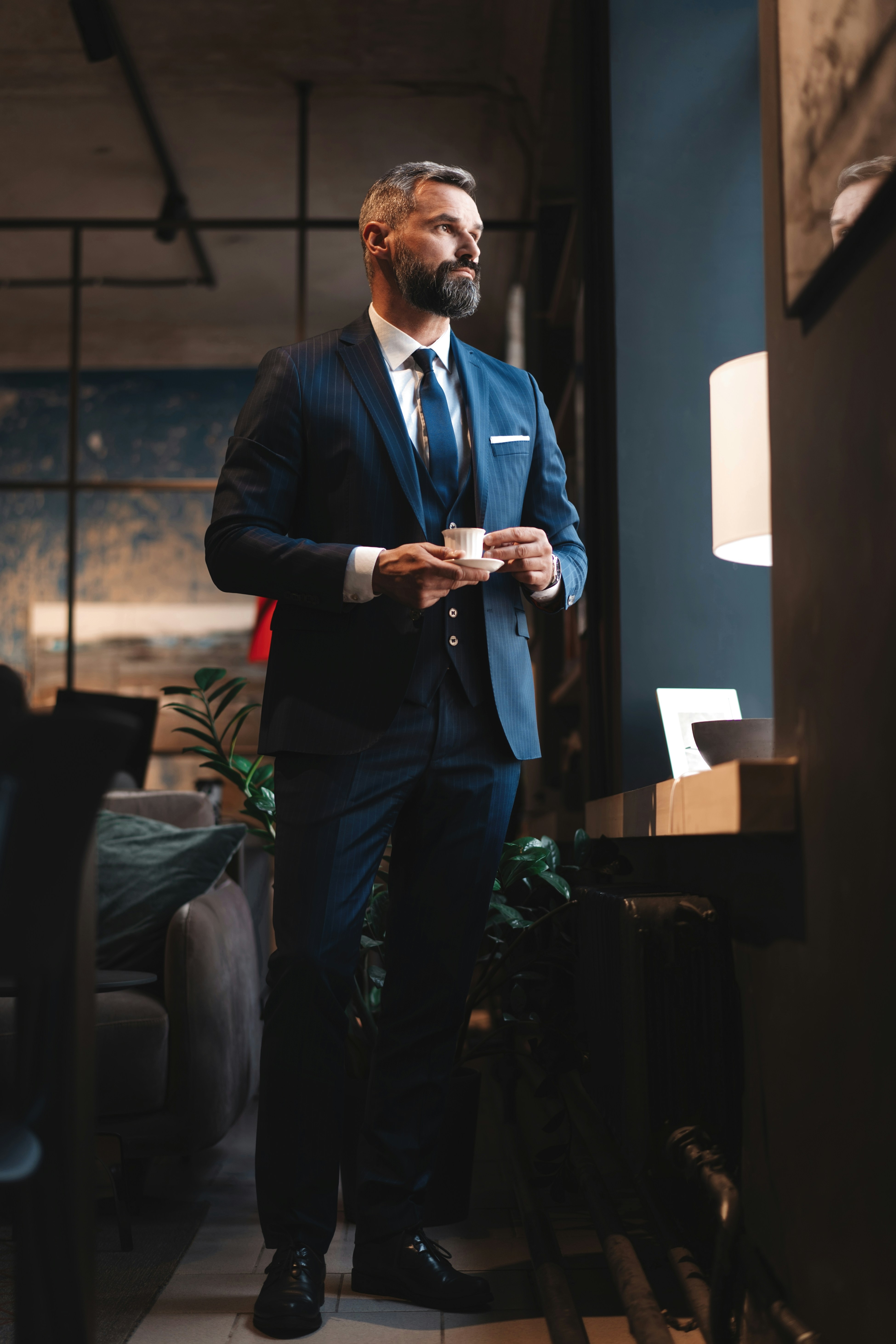 A smiling woman with long brown hair wearing a white blouse sits at a desk in a bright, modern office space, working on a silver laptop. Large windows in the background let in natural light, and a few green plants add a touch of freshness to the scene
