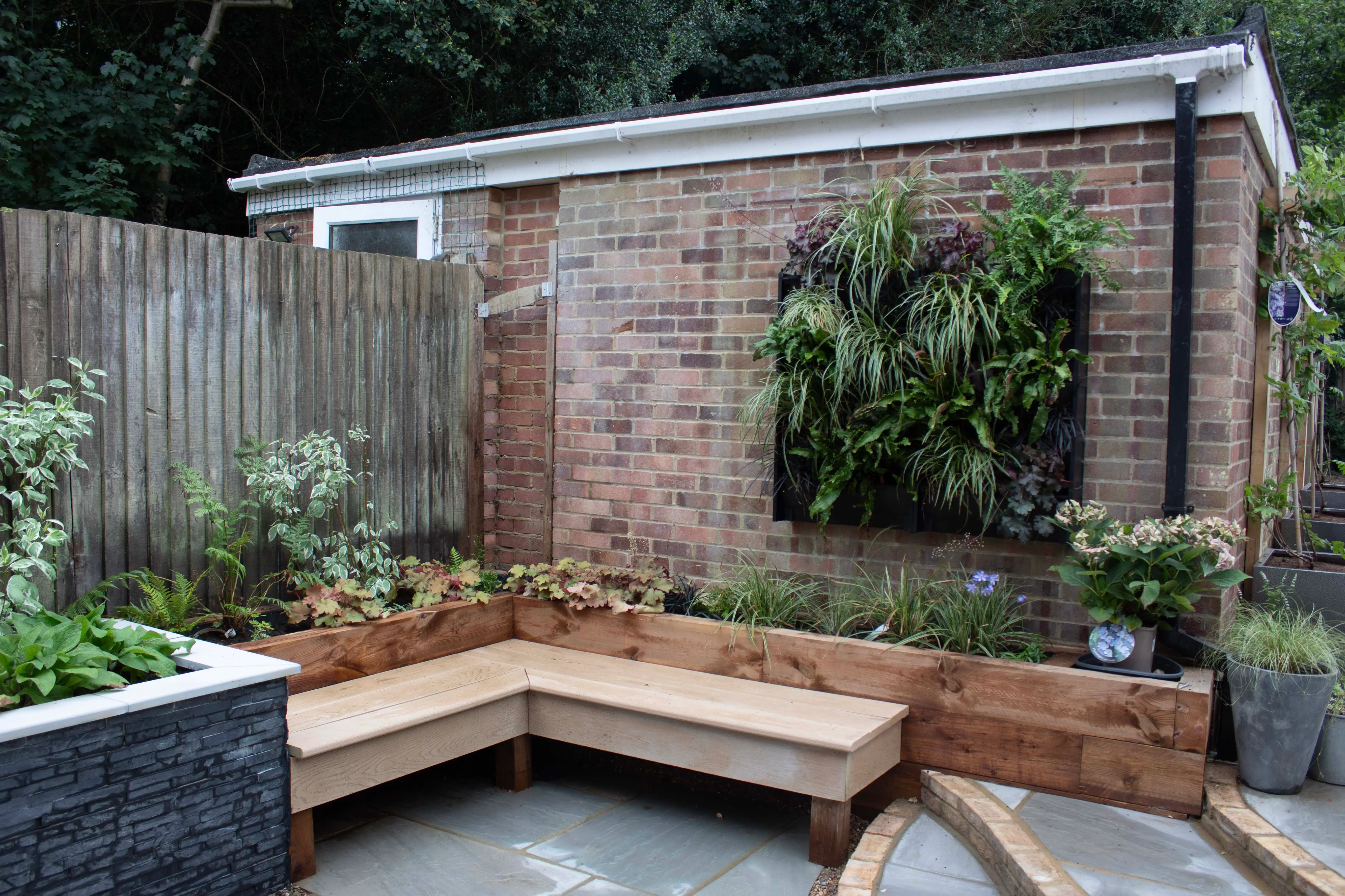 A cozy outdoor seating area with wooden benches, surrounded by greenery and a brick wall backdrop.