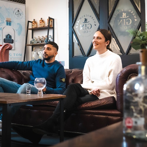 Ein Mann und eine Frau sitzen auf einem Ledersofa in einem gemütlichen Raum mit Gin-Flaschen in den Regalen und dekorativen Glastüren.