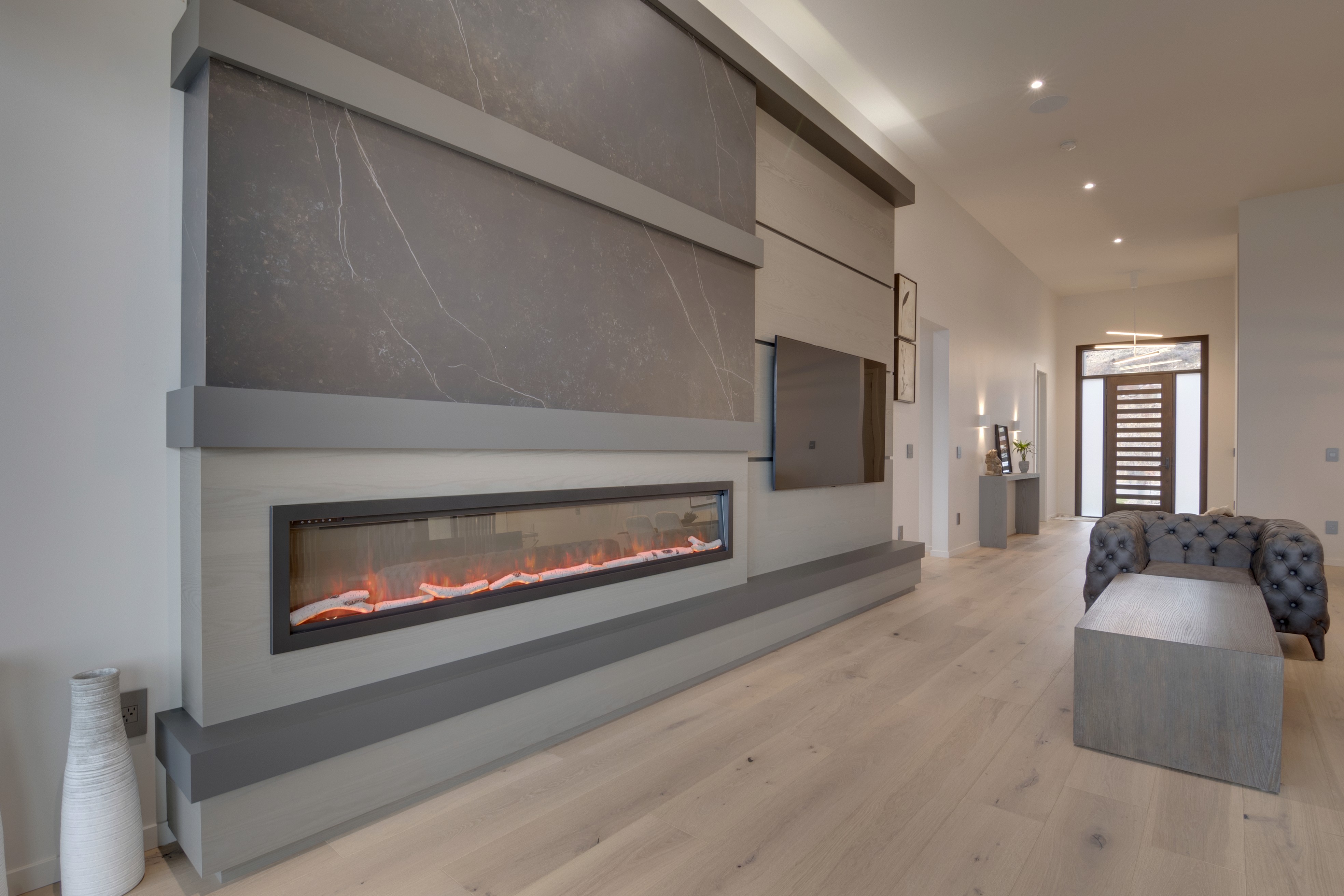Contemporary living room with custom wall paneling and built-in fireplace, featuring grey cabinetry and light hardwood flooring.