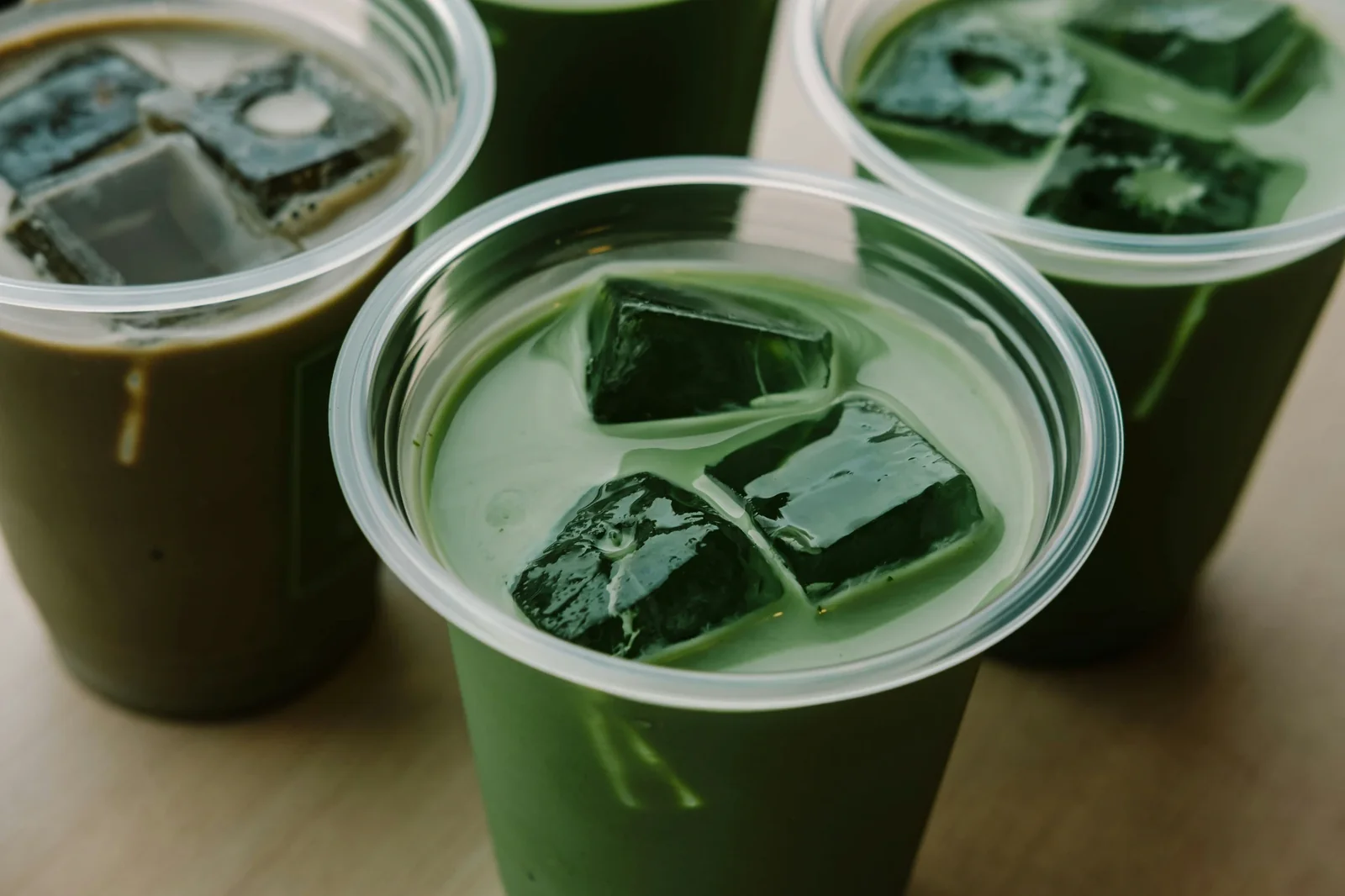 Multiple clear plastic cups containing green matcha liquid with dark matcha powder pieces floating on surface.