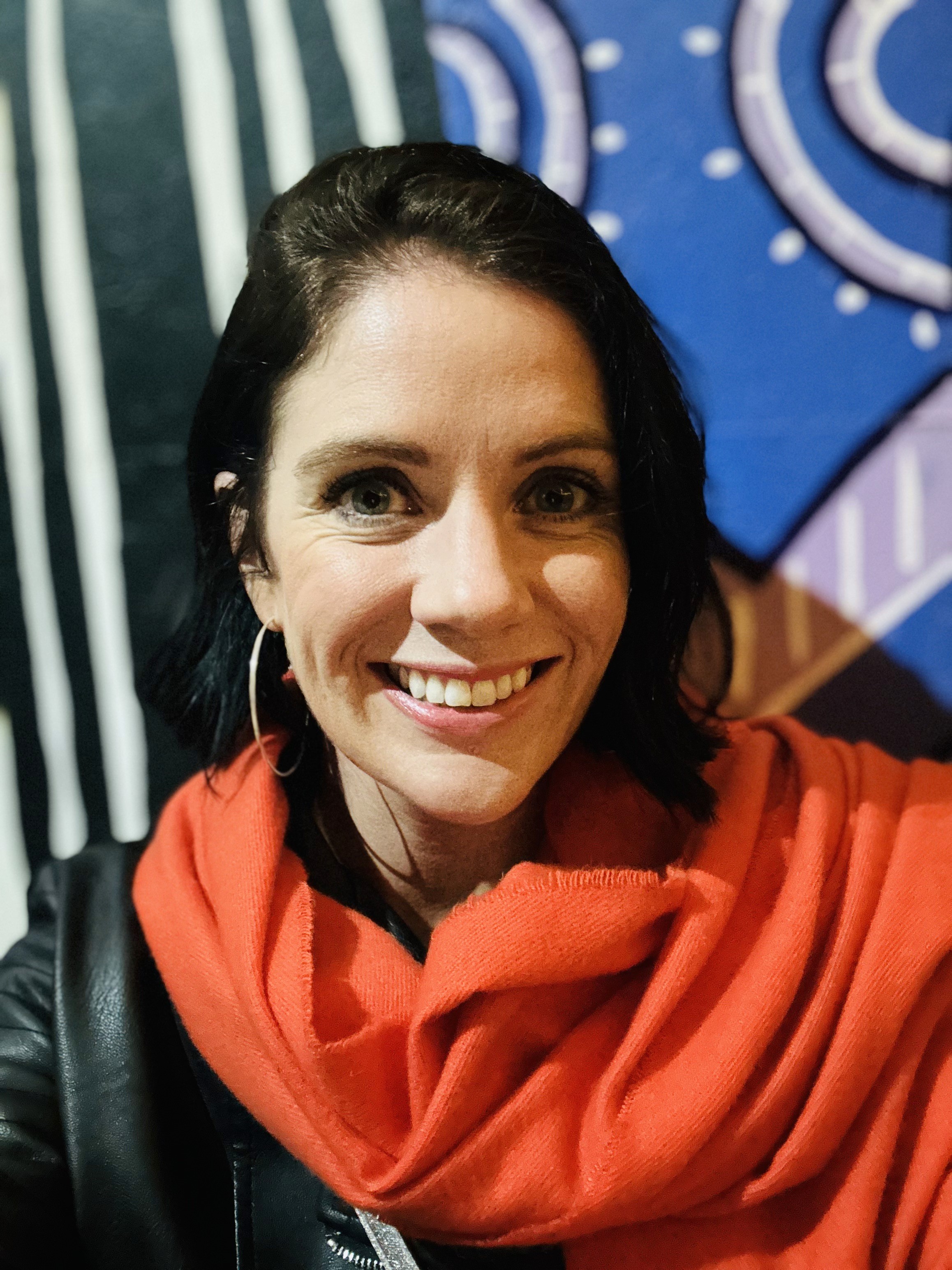 Photo of Ciara Slack, who is smiling at the camera in front of a colourful backdrop and wearing a birght orange scarf
