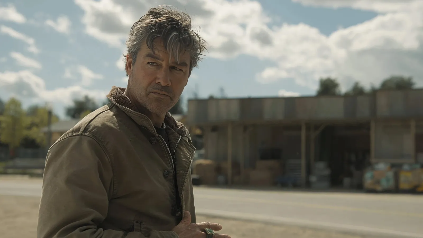 A man in a brown jacket stands on a deserted street, looking thoughtful in front of a rustic building under a partly cloudy blue sky.